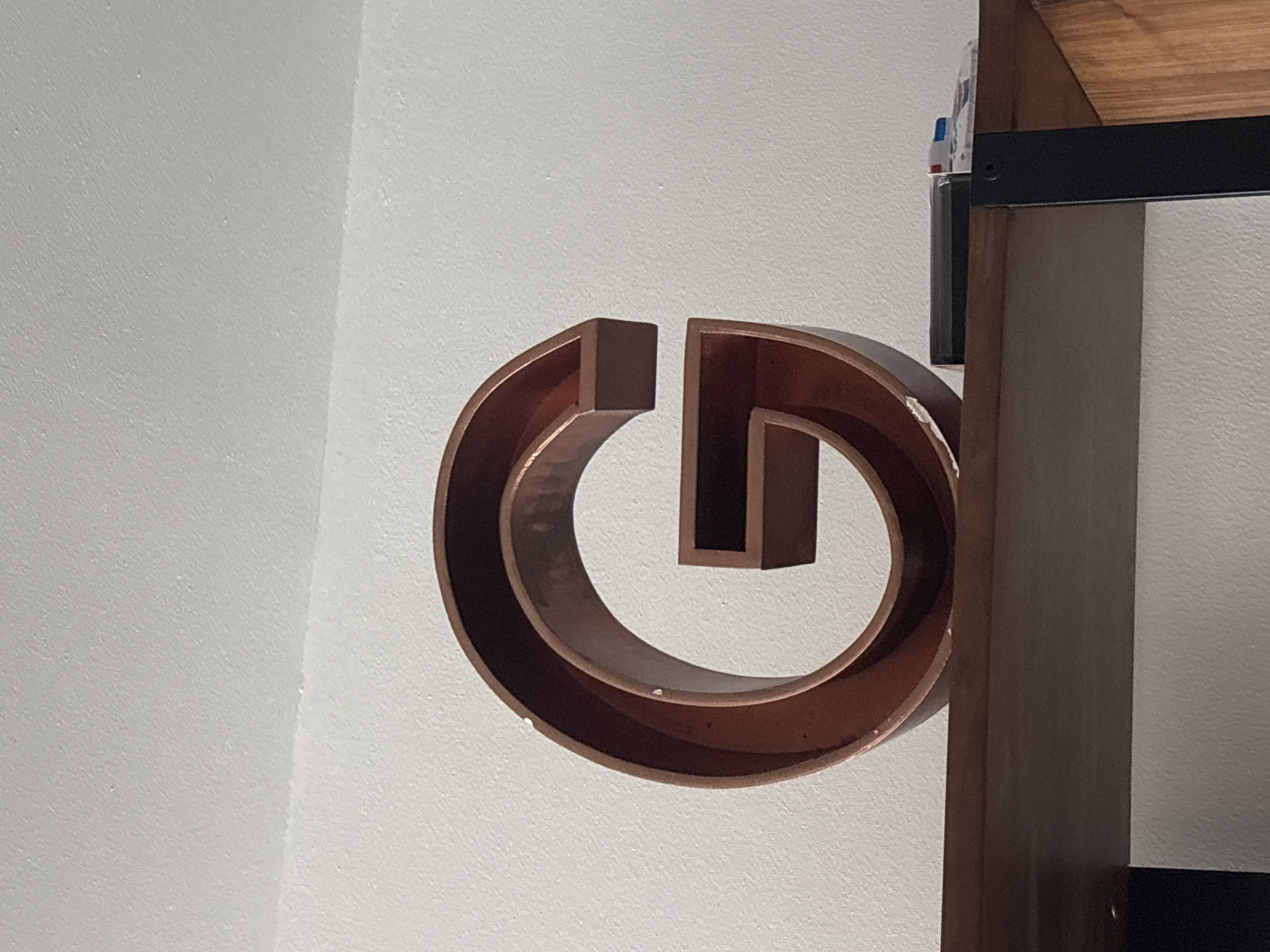 A large, reddish-brown, three-dimensional letter "G" sits on a wooden shelf against a white wall. The letter has a metallic sheen and appears to be made of wood or metal. There are some small white chips on the surface of the letter. To the right of the letter on the shelf, there are a few objects, including a black rectangular item and a small white and red object that resembles a toy car or a pen. The shelf is part of a larger piece of furniture with black metal supports and wooden accents, visible on the right side of the image. The ceiling is visible at the top of the image, with a subtle texture and a visible seam where it meets the wall. The scene is indoors, likely in a residential or office setting, and there is no indication of its location in Dubai, United Arab Emirates.