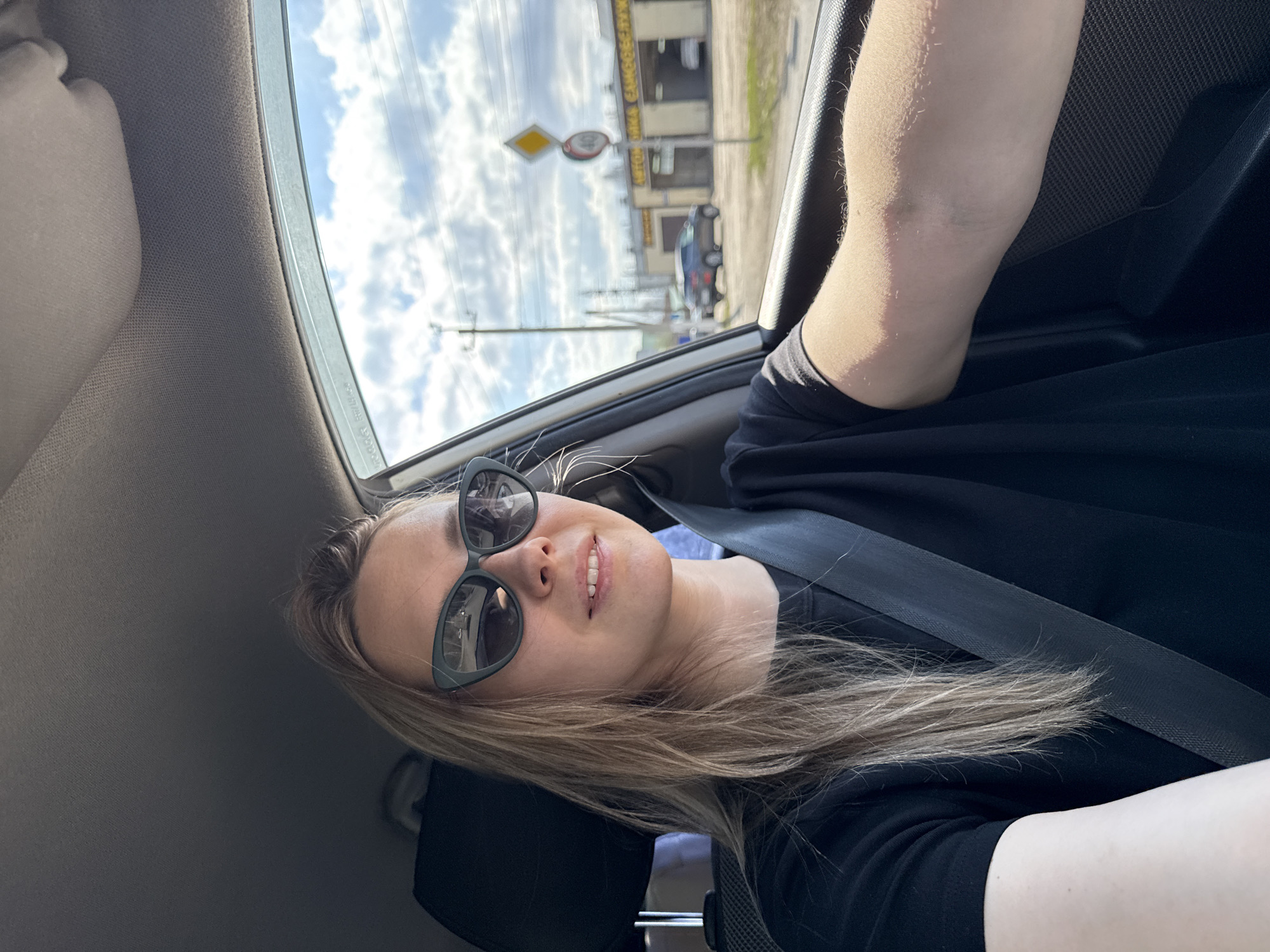 A blonde woman with sunglasses, wearing a black shirt and a seatbelt, is depicted in a selfie taken inside a car. She has a slight smile, and her head is tilted. Her hair flows over her shoulder and arm, which is resting beside her.
Through the car window, a bright, cloudy sky is visible. The car appears to be driving or parked alongside a road in what looks like an urban or semi-urban area. In the background, there is a commercial building with multiple bay doors. Visible on the building is text in Cyrillic, which reads "АВТОМОЙКА САМООБСЛУЖИВАНИЯ," translating to "SELF-SERVICE CAR WASH."
Further down the road, another car, possibly a dark SUV or hatchback, can be seen. Utility poles and power lines crisscross the sky. A yellow diamond-shaped road sign is prominent, featuring a red circle with a black '40' crossed by a diagonal line, indicating a speed limit or prohibition, with another yellow diamond sign underneath. The general setting suggests a location like Chaikovsky, Russia, consistent with the Cyrillic text and road signs. The time of day is clearly daytime, given the bright illumination.