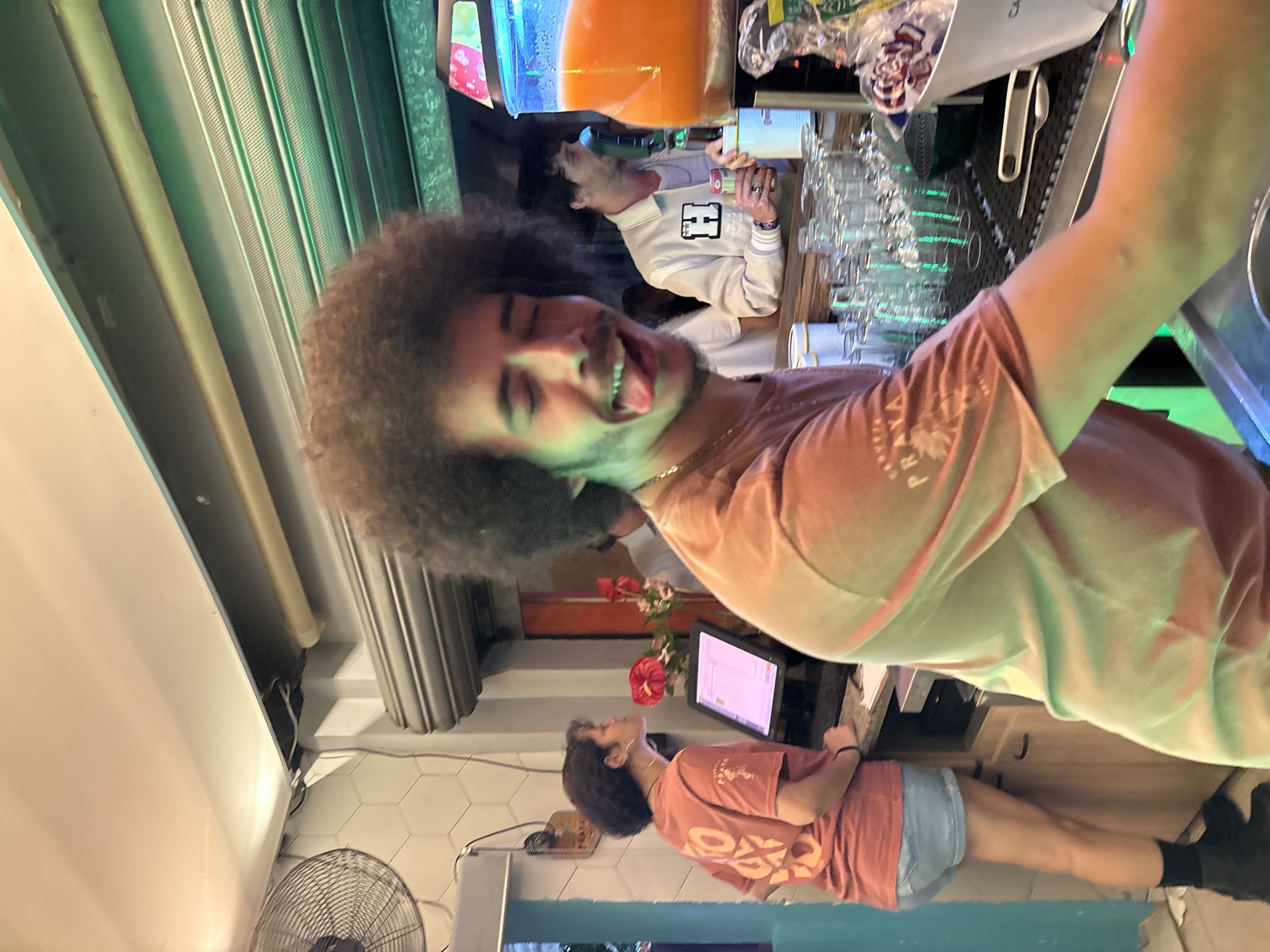 A tilted, indoor shot depicts a male with an afro sticking his tongue out for the camera. He wears a tan shirt and gold chain. Behind him, another male in a white jacket stands near a drink dispenser, likely working as a bartender. A counter with numerous glasses is present. A third male stands further in the background wearing a similar shirt to the first and is near a computer. A floor fan and tile flooring are visible in the background. The location is likely a bar in São Paulo, Brazil.