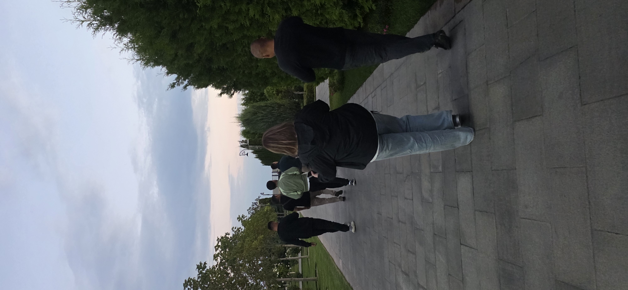 A group of six individuals, five male and one female, are walking on a paved pathway in an outdoor park. The sky above is a soft blend of blue and pink hues, indicating either dawn or dusk. The pathway is lined with well-maintained green grass and trees on both sides. On the right side, a dense row of coniferous trees forms a natural border. On the left, deciduous trees are spaced along the path, with lampposts visible further down. The individuals are walking away from the camera, towards the background where more trees and a hint of a structure can be seen. The woman in the foreground is wearing a black jacket and light blue jeans, while the men are dressed in casual attire. The scene suggests a leisurely walk in a park, possibly in the city of Cholpon-Ata, Kyrgyzstan, given the vegetation and park-like setting common in such locations.