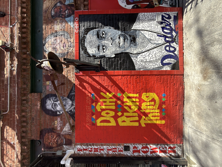 An exterior view depicts a detailed, multi-part mural on a reddish-brown brick building wall in Bedford-Stuyvesant, Brooklyn, New York City, illuminated by direct sunlight. On the upper section of the brick wall, four painted portraits are visible: an older Black man with the text "PAUL BENJAMIN R.I.P." to the far left; an older bearded Black man with "BILL LEE REST IN YOUR MUSIC PARADISE" next to him; a partially obscured older white woman; and an older Black man wearing a hat with "PLAYOFF OSSIE DAVIS REST IN PARADISE" on the far right.
Below these, a large, rectangular red panel displays the yellow text "A SPIKE LEE JOINT Do The Right Thing," accented with colorful geometric patterns. To the immediate right of this panel, and framed by red tiles, is a mosaic portrait of a young Black man, depicted from the chest up, wearing a white baseball jersey with "Dodgers" written in blue across the chest. The name "cero" is visible in black text above his head.
A white security camera is mounted on the wall's left side. An antique-style metal bell, attached to a metal bracket, protrudes from the wall above the red mural panel, partially obscuring the upper painted portraits. Vertically oriented white text reading "FIGHT THE POWER" is painted on a red strip to the left of the "Do The Right Thing" panel. A concrete sidewalk is visible at the base of the wall, featuring shadows cast by the building or nearby structures.