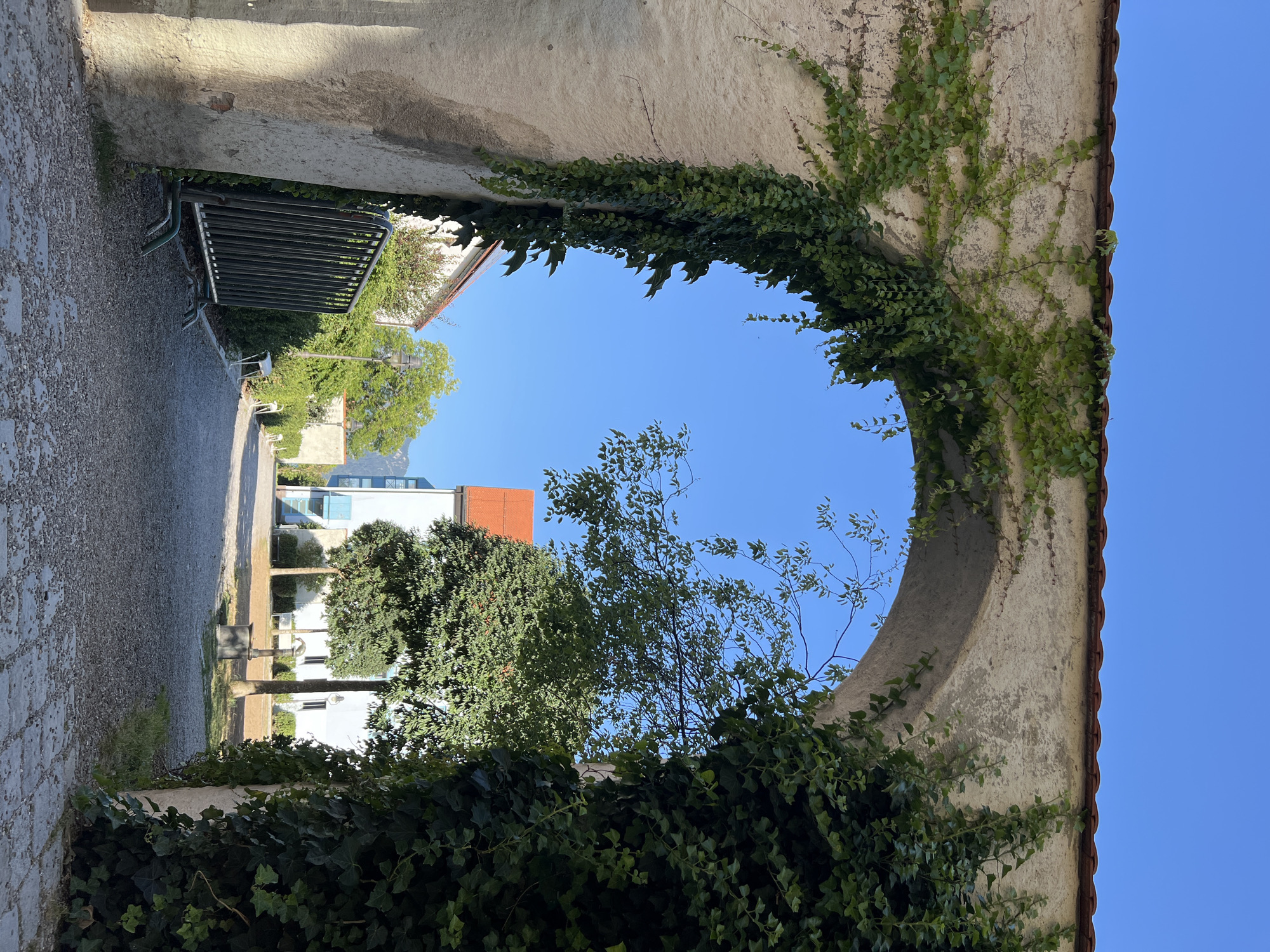 An arched passageway covered in ivy leads to a courtyard. The passageway is constructed of weathered stone and topped with red tiles. In the courtyard, a gravel path winds past trees and buildings. One building has a red roof, another is white with blue accents. A metal fence is visible to the left of the passageway. The sky is clear and blue. The scene appears to be in Immenstadt im Allgäu, Germany, given the architectural style and landscaping.