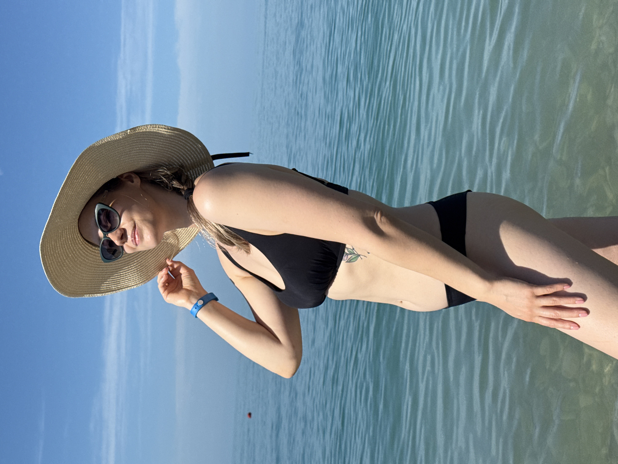 A woman stands in the shallow, clear waters of the Black Sea, off the coast of Nebug, Russia, under a bright blue sky with a few wispy clouds. She is presented in a three-quarter profile, looking upwards and to her right, with a slight smile. She has blonde hair, partially braided, and wears a wide-brimmed straw hat and cat-eye sunglasses with dark lenses and light-colored frames. Her attire consists of a black two-piece bikini. A blue wristband is visible on her left wrist, which is raised with her hand gently touching the brim of her hat. A small, green, floral-patterned tattoo is visible on her left ribcage. The water around her is calm, displaying varying shades of turquoise and blue with visible ripples, and a faint red spherical buoy is situated in the mid-ground to her left.