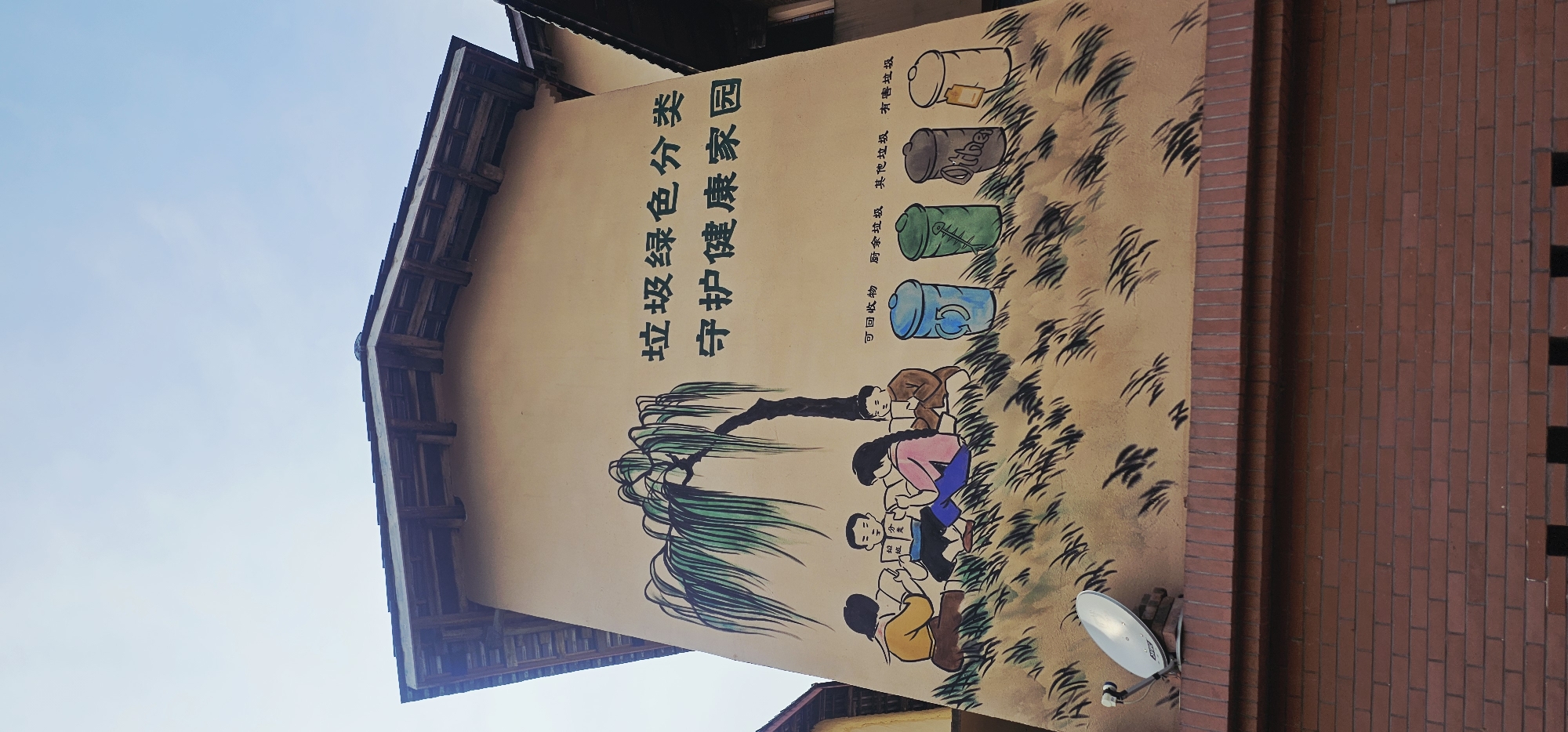 The image depicts a mural painted on the exterior wall of a building in Shapingba District, China. The mural features a weeping willow tree with its branches cascading down. Beneath the tree, four children are seated on the ground, each engrossed in reading books. To the right of the children, five distinct trash cans are painted, representing different categories of waste. Above the mural, Chinese characters are inscribed, conveying a message about "garbage green classification" and "protecting a healthy home." The building has a tiled roof and a brick base. A satellite dish is mounted on the lower left portion of the wall. The sky above is a pale blue, suggesting daytime.