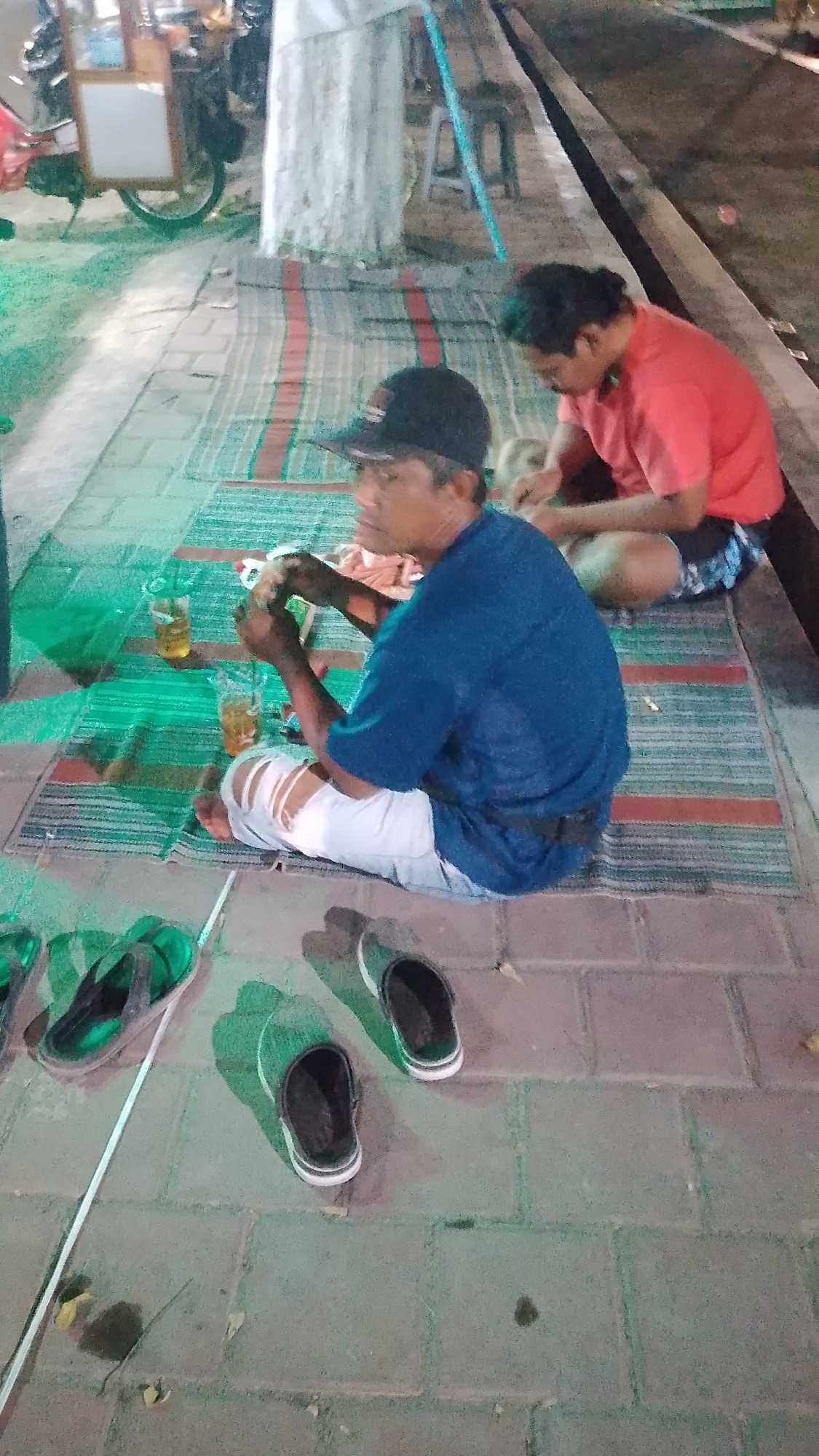 In the city of Karanglangu, Indonesia, two individuals are observed sitting on mats laid out on a paved walkway. The person in the foreground, wearing a blue shirt and ripped white pants, is looking down at their hands, which appear to be holding food or an object. To their right, a person in a red shirt is also looking downwards, seemingly engaged in an activity. Two glasses containing a yellowish liquid are visible on the mat near the person in the blue shirt. In the background, a food cart is partially visible. Several pairs of footwear, including flip-flops and slip-on shoes, are scattered on the ground around the individuals. The scene is illuminated by artificial lighting, casting shadows and creating a slightly blurred effect. The ground is paved with rectangular bricks, and there are some fallen leaves.