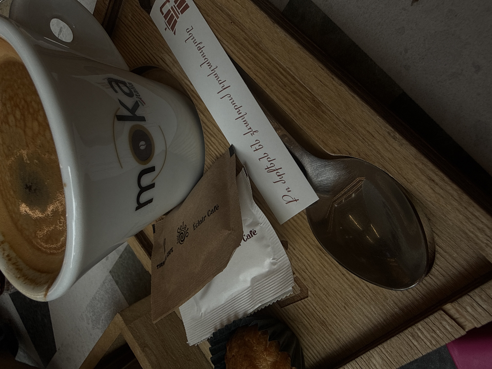 A top-down, canted, close-up shot shows a white "moka" coffee cup filled with coffee on a wooden tray. Next to the cup is a brown paper sugar packet from "Eclair Cafe" and a white sugar packet. A silver spoon lies adjacent to the packets, with a white paper slip imprinted with Armenian script resting on the spoon. A partially visible pastry in a black paper cup is located at the bottom of the frame. The scene is indicative of a coffee setting in Yerevan, Armenia, given the Armenian script and the Eclair Cafe branding. A gray patterned surface is visible beside the tray on the lower-left side of the frame.