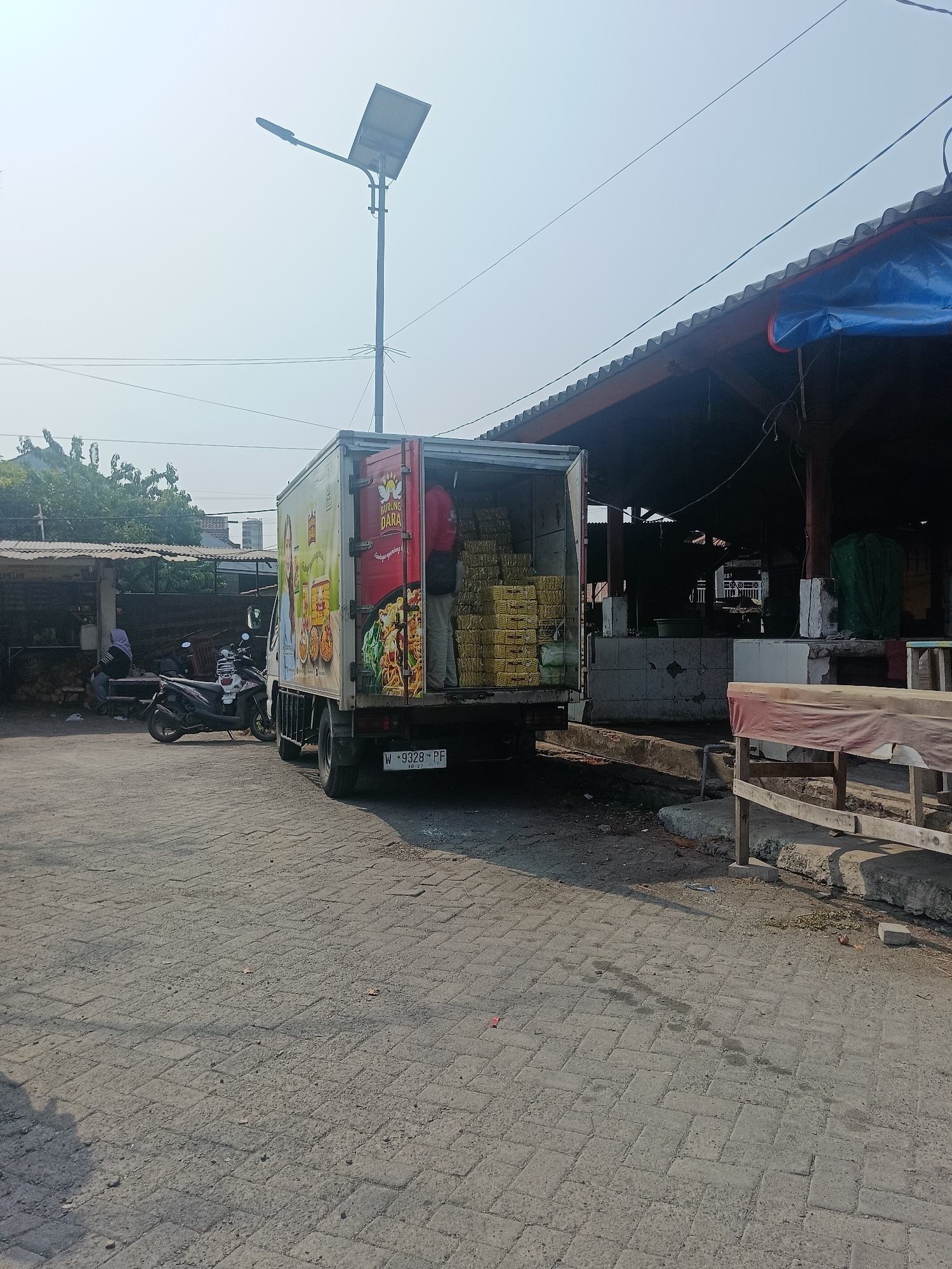 This is a daytime, eye-level, medium shot of a street scene in the city of Ketajen, Indonesia. A delivery truck is parked at an outdoor market. The truck is loaded with boxes of noodles. A person in a red shirt stands inside the back of the truck. A motorcycle is parked in front of a market stall. A solar-powered street light is visible in the background. The ground is paved with interlocking bricks.