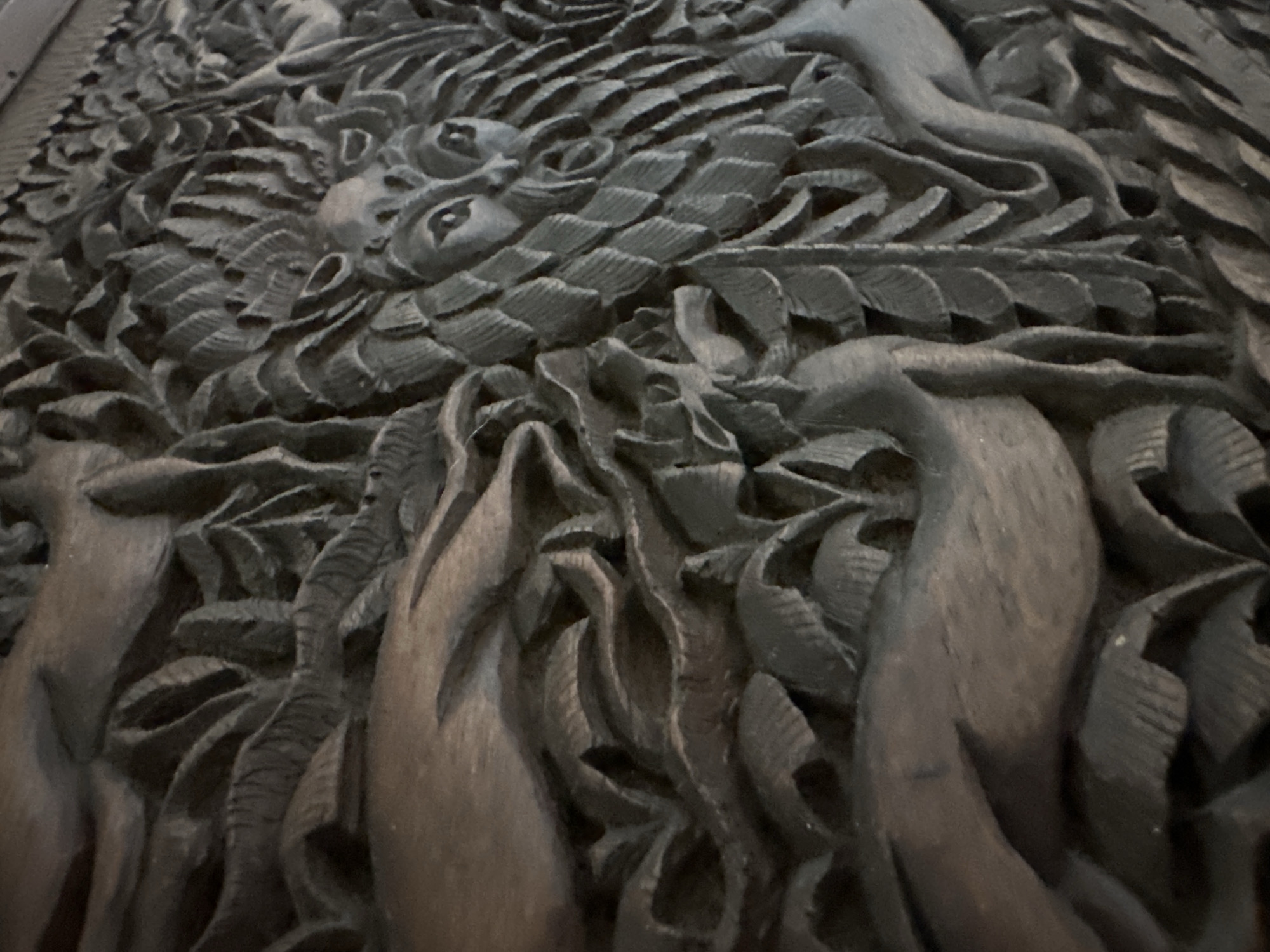 The image shows a detailed wood carving. The carving depicts a scene with a face with prominent eyes and a leafy headdress, possibly representing a nature spirit or deity. To the left of the face, a curved form suggestive of an animal's body, perhaps a deer or mythical creature, is visible, with a leg extending downwards. To the right of the face, more leaves and foliage are carved, creating a textured background. The carving has a dark, weathered appearance, indicating it is made of wood. There is no discernible location information within the image, and therefore it cannot be placed in Thora, Australia.