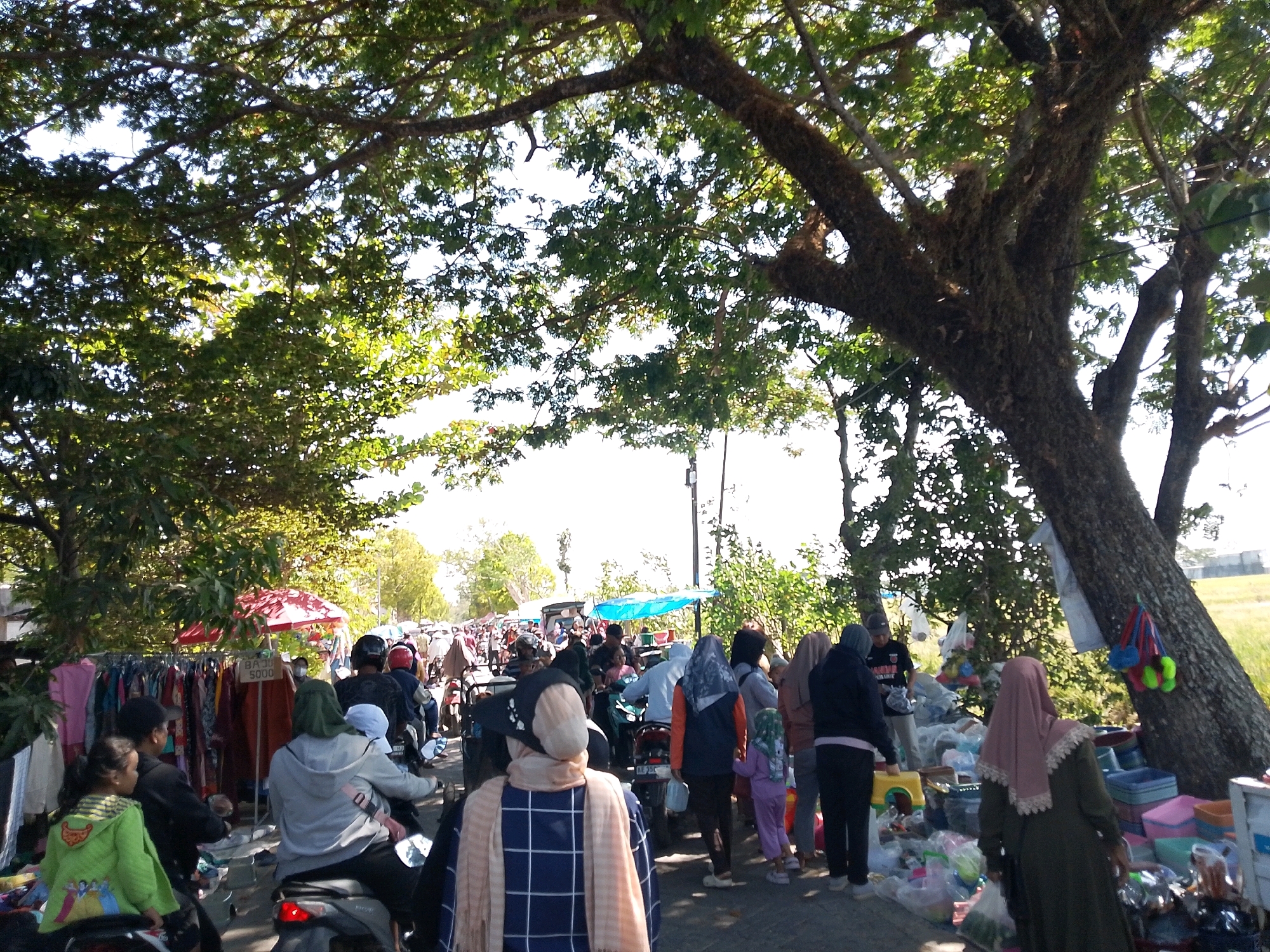 The image depicts a bustling street market scene under the shade of large trees in Tepas, Indonesia. In the foreground, a young girl in a green hooded jacket with character prints sits on a motorcycle. Next to her, a person in a black cap is also on a motorcycle. Further to the left, a rack of colorful clothing is displayed for sale, with a red umbrella providing shade. A sign on the clothing rack reads "BAJU 5000". In the midground, several people are seen on motorcycles, some carrying passengers. One person wears a grey hooded jacket and is carrying a child. Another individual is wearing an orange jacket and a patterned headscarf. The market extends into the background with more people, stalls, and motorcycles. The environment is characterized by a road lined with trees, suggesting an outdoor market setting. The lighting indicates it is daytime. To the right of the image, a large tree with thick branches dominates the scene, with some items, possibly toys or decorations, hanging from its trunk. Several plastic containers and other items are piled up near the base of this tree, suggesting more merchandise. The overall atmosphere is one of activity and commerce in a local market.