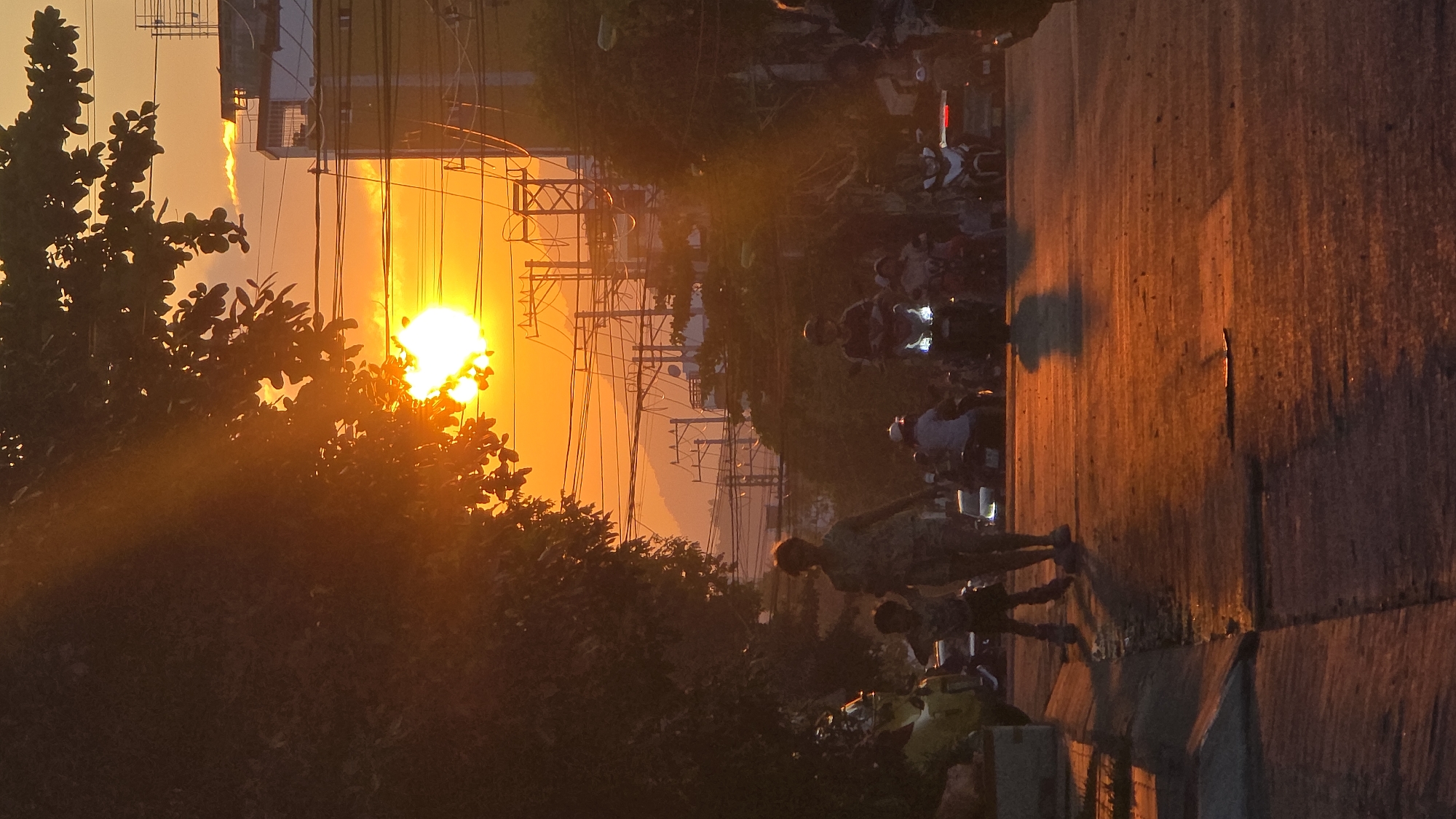 The image depicts an urban street scene in Nha Trang, Vietnam, captured during either sunrise or sunset, characterized by intense backlighting. A large, radiant orange sun is prominently visible in the upper left, partially obscured by dark, silhouetted tree foliage. The sky exhibits vibrant orange and yellow hues, transitioning to lighter tones. A dense network of black overhead power lines and utility poles spans the upper half of the frame, with layered, dark mountains visible in the distant background against the horizon.
On the paved asphalt road in the foreground, an adult and a small child, both silhouetted, walk away from the camera on the left side. Further down the street, multiple motorbikes with riders, some illuminated by bright white headlights and others appearing as dark silhouettes, are present, alongside several indistinct cars. One motorbike with a rider wearing a helmet is notably visible in the mid-ground. The road surface and the curb on the left are bathed in an orange glow, revealing texture and casting faint shadows towards the viewer. Buildings are discernible in the mid-ground and background, adding to the urban environment.