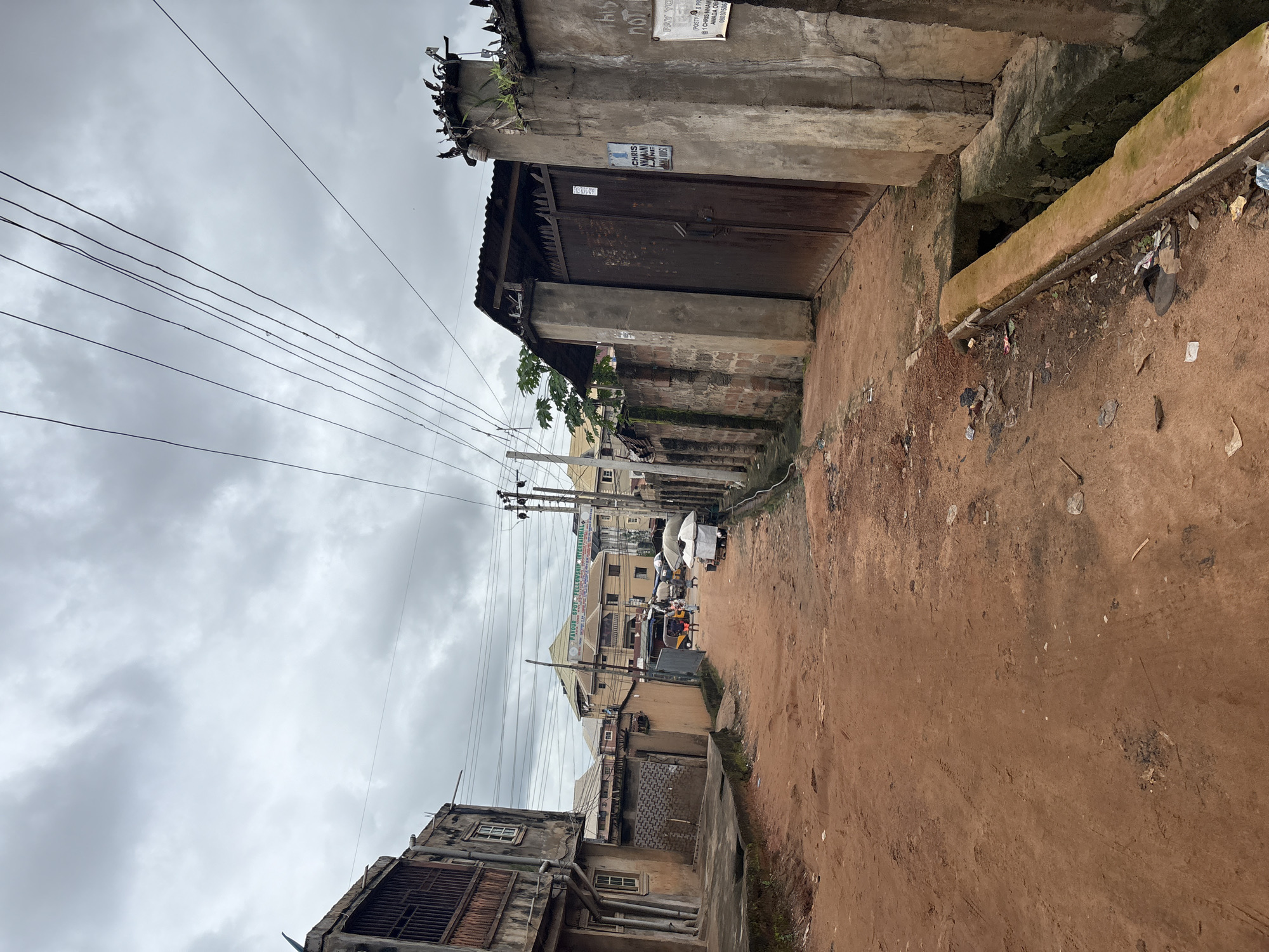 This is a street-level view of a red dirt road in Nkpor, Nigeria. The road is lined with buildings and a large, rusted metal gate. Overhead, power lines crisscross the cloudy sky.
On the left side of the road, a two-story building shows signs of wear, with a barred window on the upper floor and a partially visible lower floor. Further down the street, more buildings are visible, including a larger, modern-looking structure with a banner that reads "FAVOUR SPOT FELLOWSHIP INTERNATIONAL." A small, three-wheeled vehicle, possibly a tuk-tuk, is stopped in the road, with several people gathered around it. A dog can be seen in the distance.
On the right side of the road, a concrete wall borders the street, topped with jagged metal spikes and sparse vegetation. A large, dark metal gate is set within this wall, with a sign that reads "1 CHRIS NKAMANI LANE." The wall also has graffiti and a sign that says "POST IT & PR...". A concrete drainage ditch runs along the edge of the road, filled with debris.
The overall impression is of a working-class urban environment. The ground is dusty and uneven, with scattered litter. The sky is overcast, suggesting a potentially humid or rainy day.