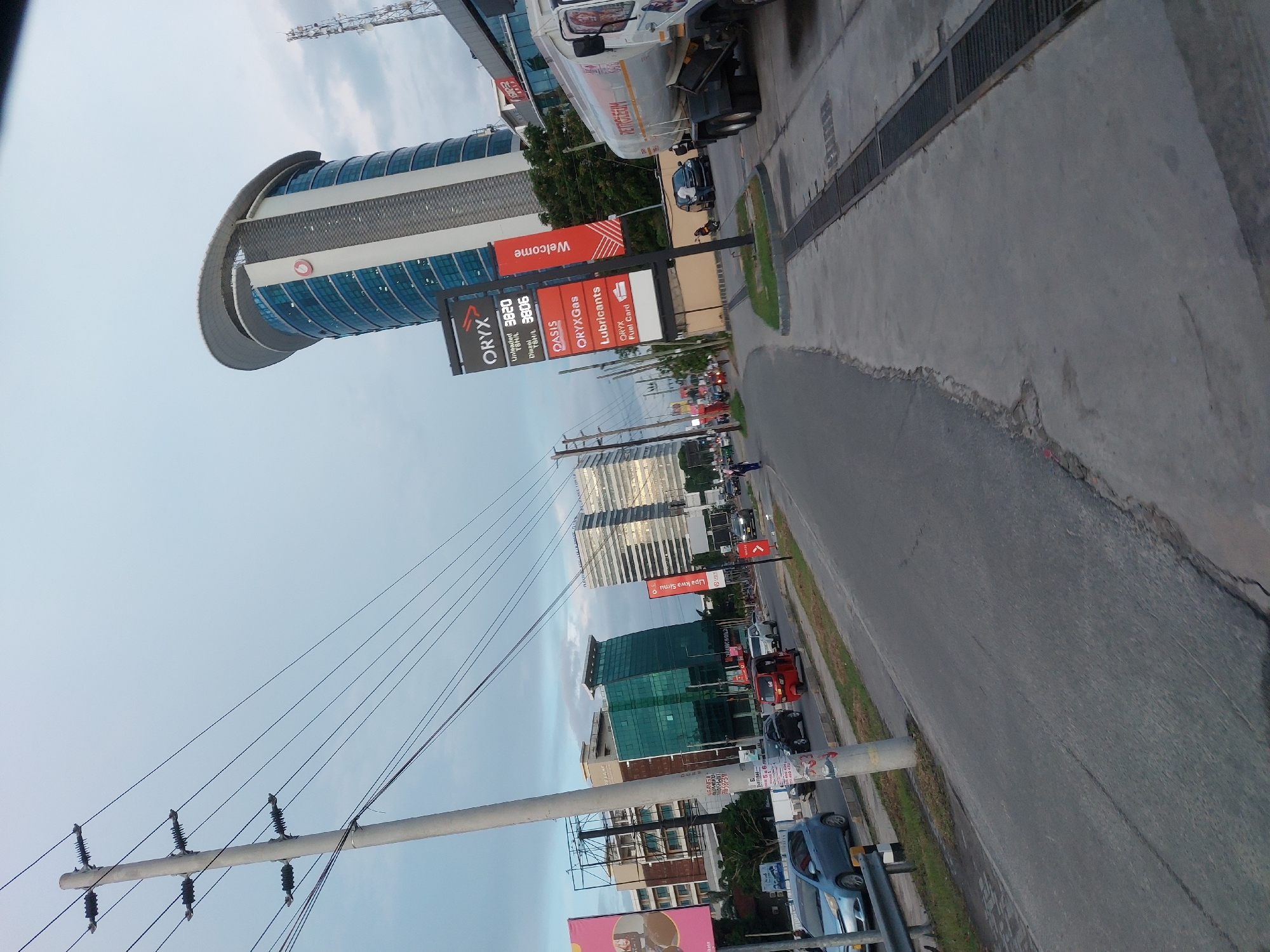 The image depicts a street scene in Dar es-Salaam, Tanzania. In the foreground, there's a view of an Oryx petrol station with a sign displaying fuel prices and advertisements for lubricants. Above the station, an elevated, contemporary curved building is visible. The street is lined with various buildings, including a modern green-glass office complex. The sky is overcast, with visible power lines running parallel to the street. Cars are parked along the side of the road, and there's moderate traffic. The focus is on the gas station and the surrounding urban architecture.