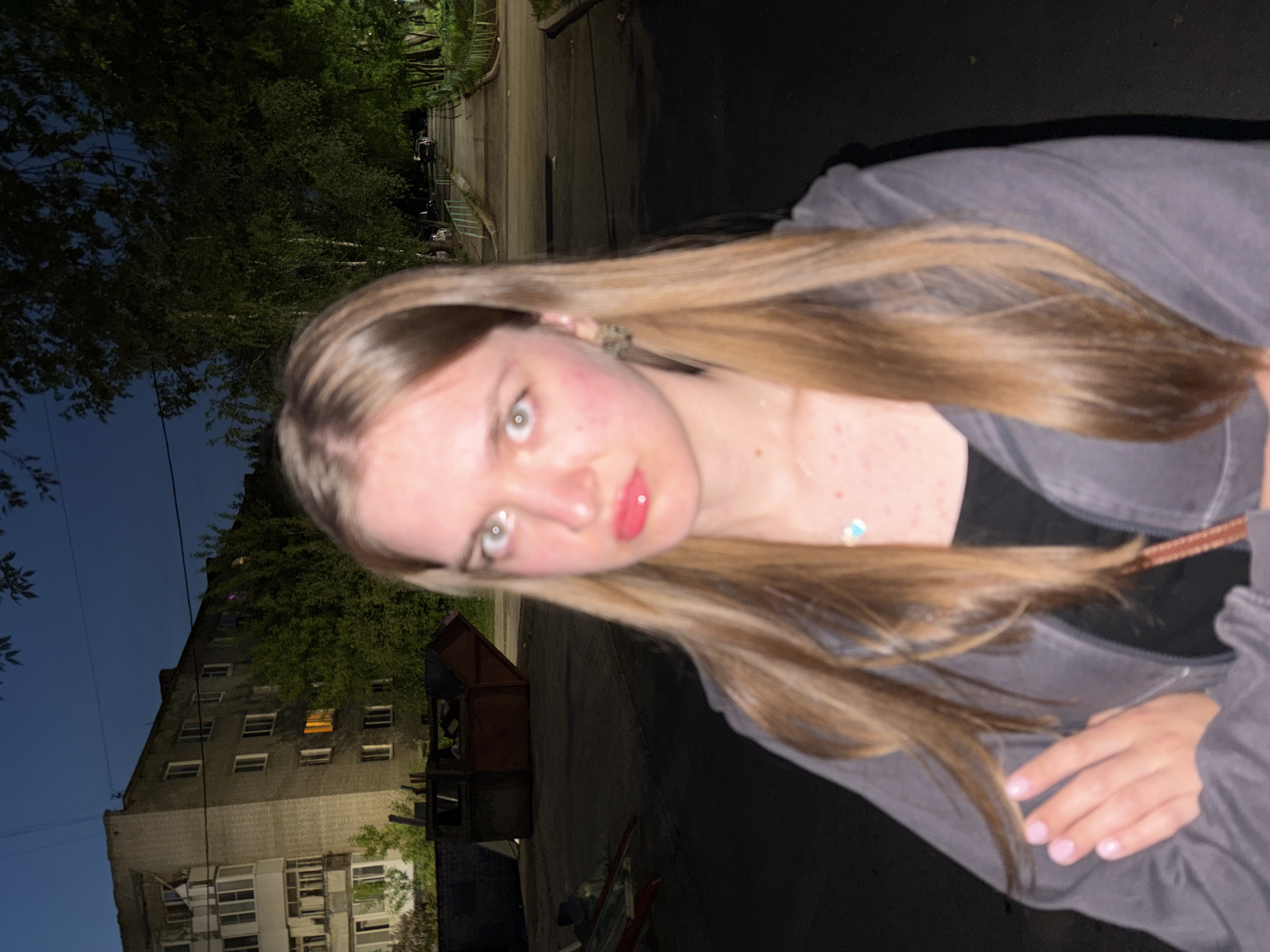 A young woman poses for a selfie at night. She has long, light brown hair and is wearing a dark gray jacket. Her lips are glossed with a pink tint. Behind her is a multi-story building with many windows, some illuminated, suggesting residents are home. The presence of trees and the overall urban setting suggest this could be in Perm, Russia. A wooden structure, possibly a playhouse or storage unit, is to the left of the building.
