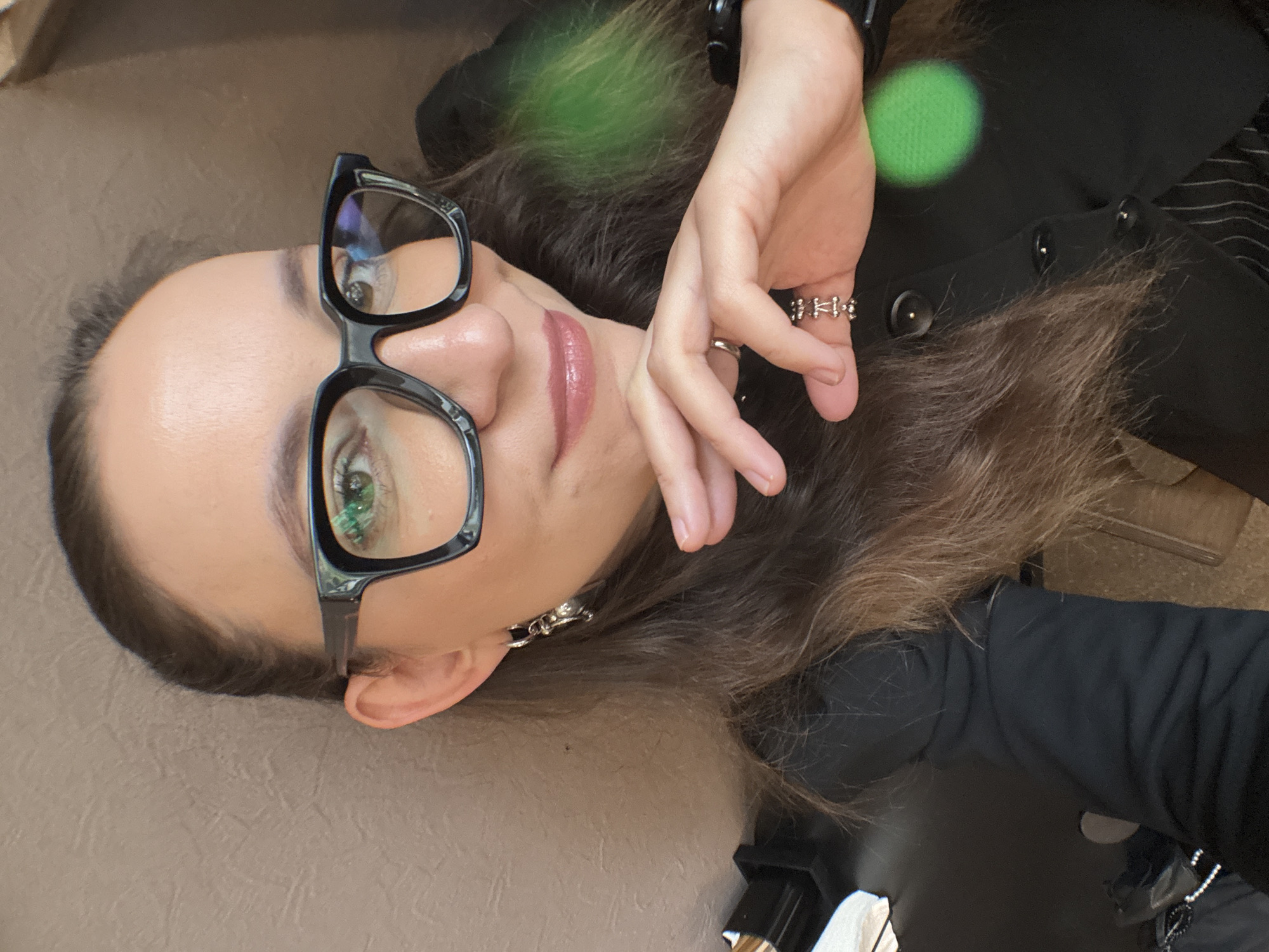 A medium close-up selfie shows a woman from the chest up, her head angled slightly to her left. She is wearing large, black-rimmed glasses and has long, brown hair with highlights. Her right hand is raised to her face, with her fingers lightly touching her cheek. She is wearing rings on two fingers of her right hand: a thin silver band on her ring finger and a more ornate silver ring on her middle finger. She is also wearing a dangling silver earring in her left ear. She has on a black collared shirt with visible buttons, and a hint of pinstripe fabric is visible on her lower chest. The background consists of a textured, neutral-colored wall to her left and a dark surface with some light reflection to her right. There is a blurred green circle in the foreground on the right side of the image, possibly a light reflection or lens flare. The overall lighting is soft. There is no indication of the city of Chaikovsky, Russia, in the image.