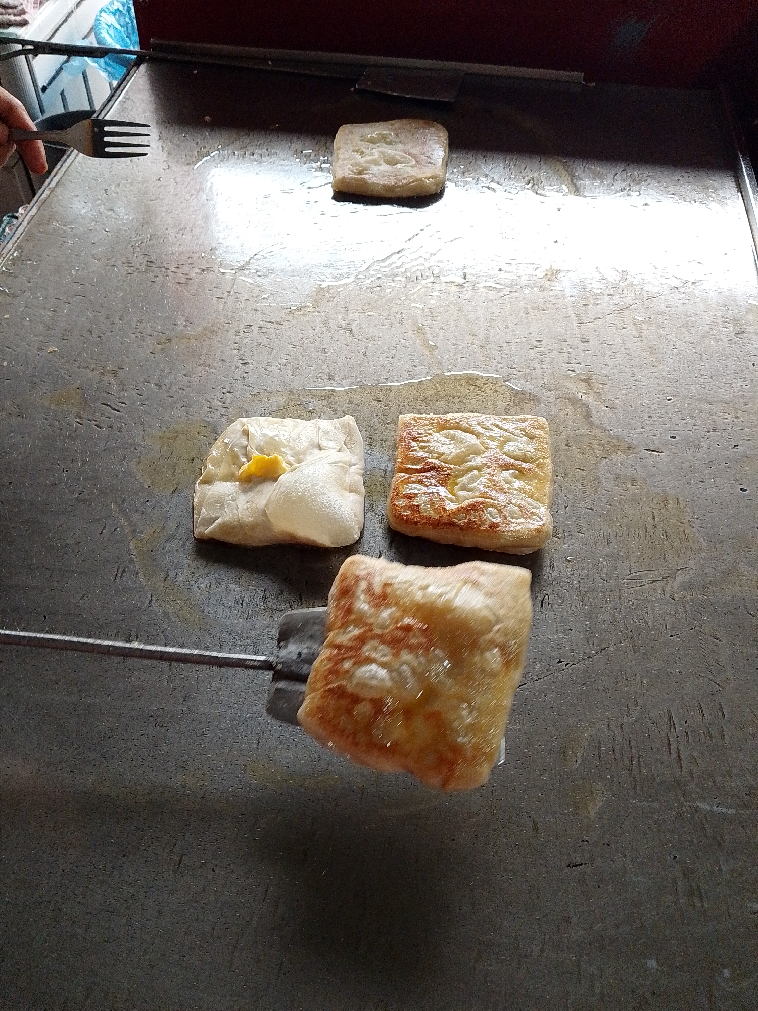The image shows four squares of food being cooked on a flat-top griddle. The griddle appears to be made of metal and has a slightly oily surface. In the background, there is a reddish-brown appliance and a blue plastic bag. A hand holding a fork is visible on the left side of the frame, suggesting someone is preparing or interacting with the food.
Three of the food squares are already on the griddle. One, in the upper center, is pale and appears to be plain. To its left, another square is partially folded, revealing a yellow filling, possibly egg or cheese. To the right of the plain square is a golden-brown square with a patterned surface, indicating it has been cooked and browned. A fourth square, held by a spatula, is in the foreground, also golden-brown and displaying a similar patterned texture. The overall impression is that these are savory pancakes or crepes being fried. The lighting suggests it is daytime, and the setting appears to be an outdoor food stall or a kitchen. There is no visible text in the image.