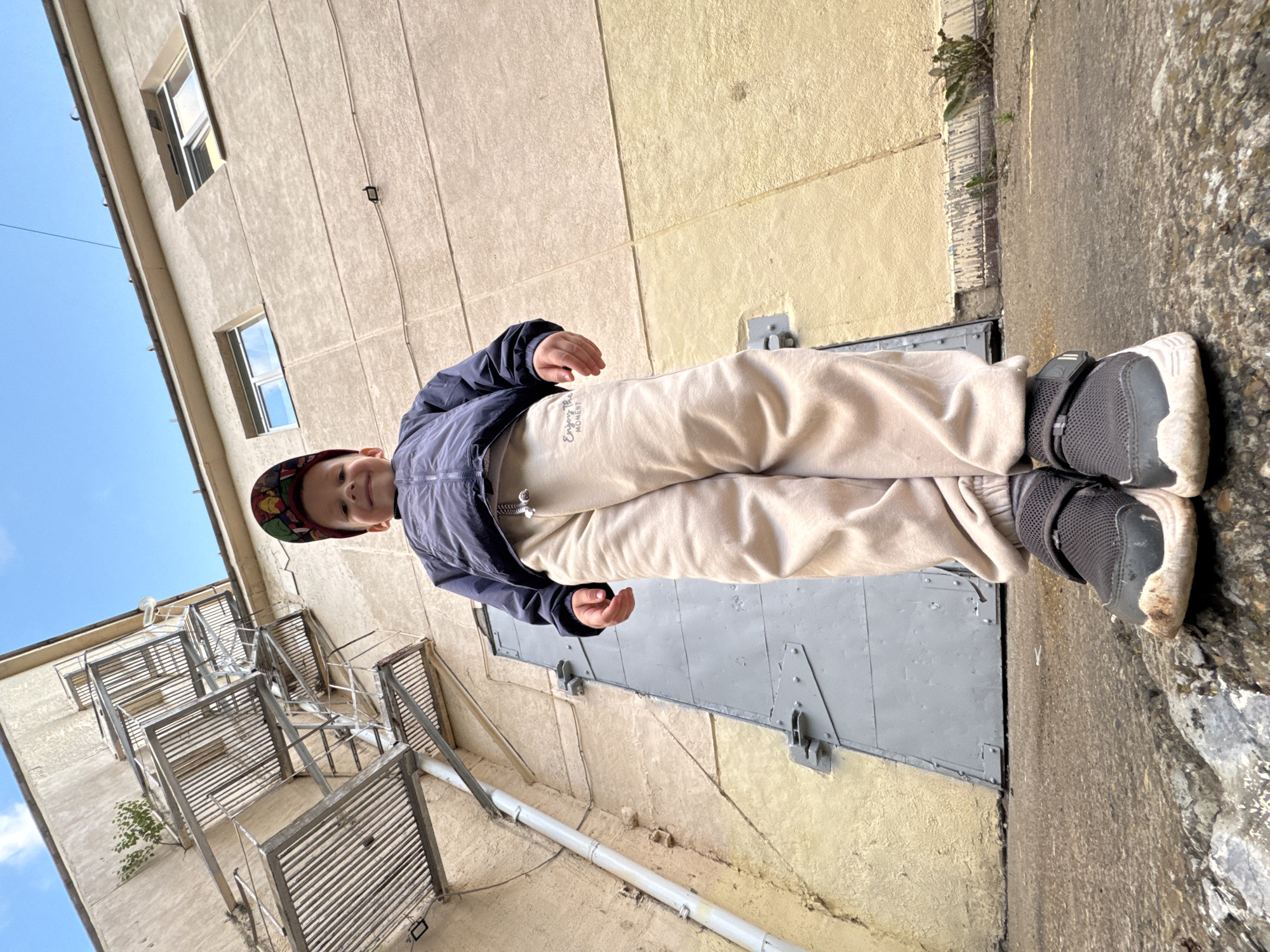 The image shows a young boy, likely a toddler, standing in an outdoor alleyway. He is wearing a dark blue jacket, light-colored pants, a patterned baseball cap, and dark sneakers. The boy is smiling and looking up at the camera, appearing happy and engaged.
The setting is an urban alley with a textured, light-colored building on one side and a rocky, uneven ground on the other. The building has several windows and an external metal fire escape. There is a large gray metal door set into the building's wall. The ground appears to be a mix of concrete and rough stone.
The time of day appears to be daytime, with clear blue skies visible. The lighting suggests it's likely sunny.
Visible text on the boy's pants reads "Enjoy The Moment". The location context is Chaikovsky, Russia.
