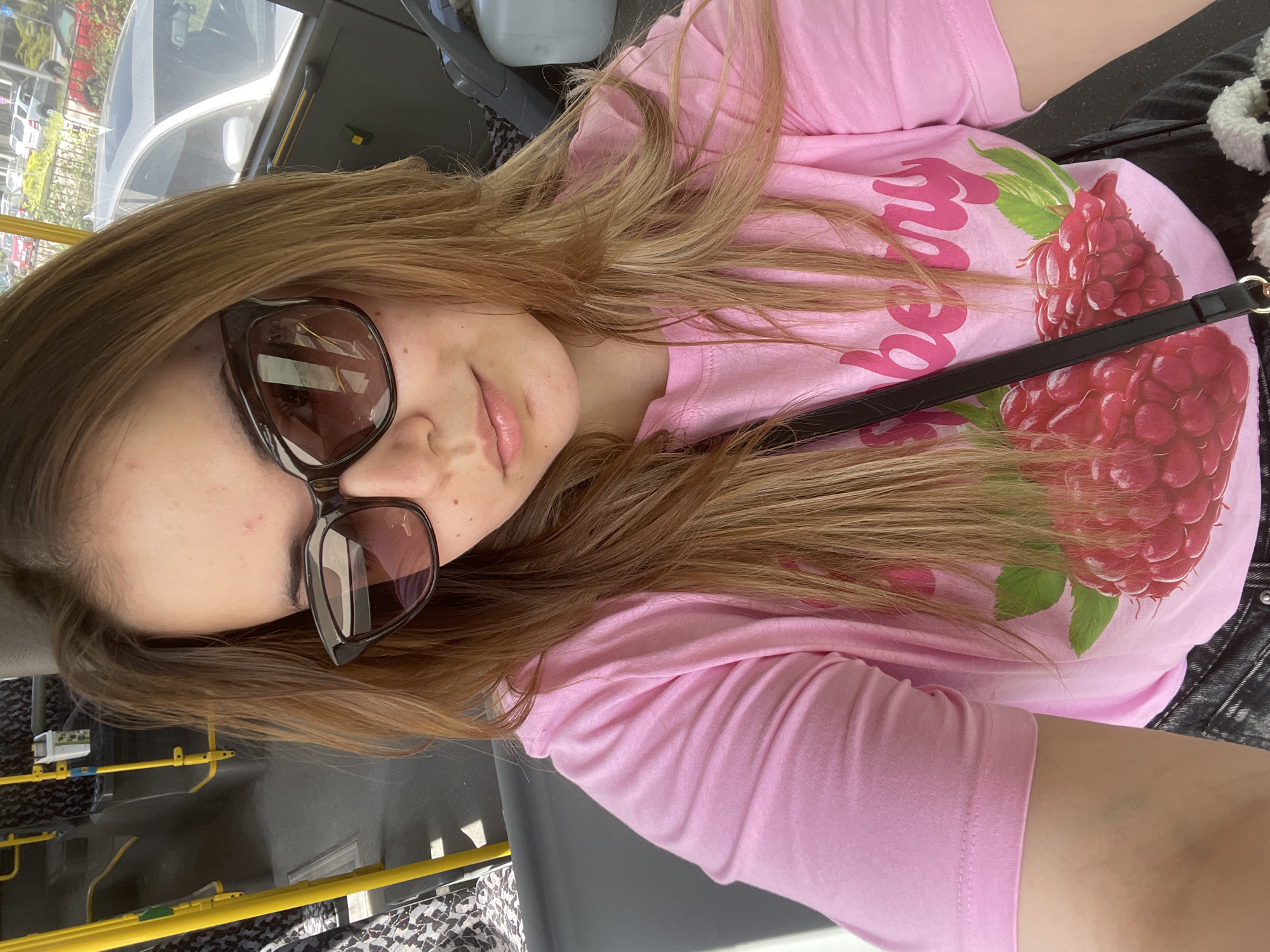 A woman with long, light brown hair is wearing sunglasses and a pink T-shirt with a raspberry design. She is sitting on a bus, possibly in the city of Plovdiv, Bulgaria, as indicated by the visible bus interior. The bus features yellow handrails and a gray seat. Outside the bus windows, there are glimpses of trees and possibly buildings, indicating an urban environment. The woman has a purse with a black strap across her body.