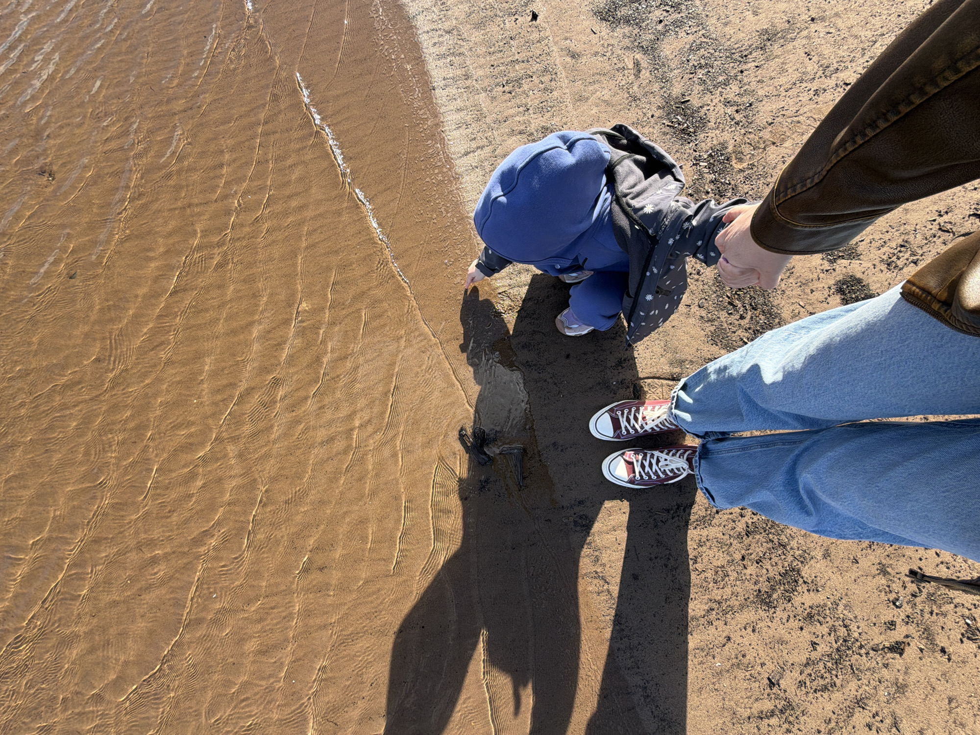 A photograph depicts a person and a toddler at the edge of a body of water with a sandy shore. The scene is likely in Noviy, Russia. The person is wearing blue jeans and maroon Converse sneakers. The toddler, dressed in a blue hooded outfit, is reaching towards the water. The person is holding the toddler's hand. The shoreline consists of sand, partially submerged by shallow water. The sand transitions from wet near the water's edge to dry further inland. The person is wearing a leather jacket. The photograph is taken from an overhead perspective, with the subjects casting shadows onto the sand.