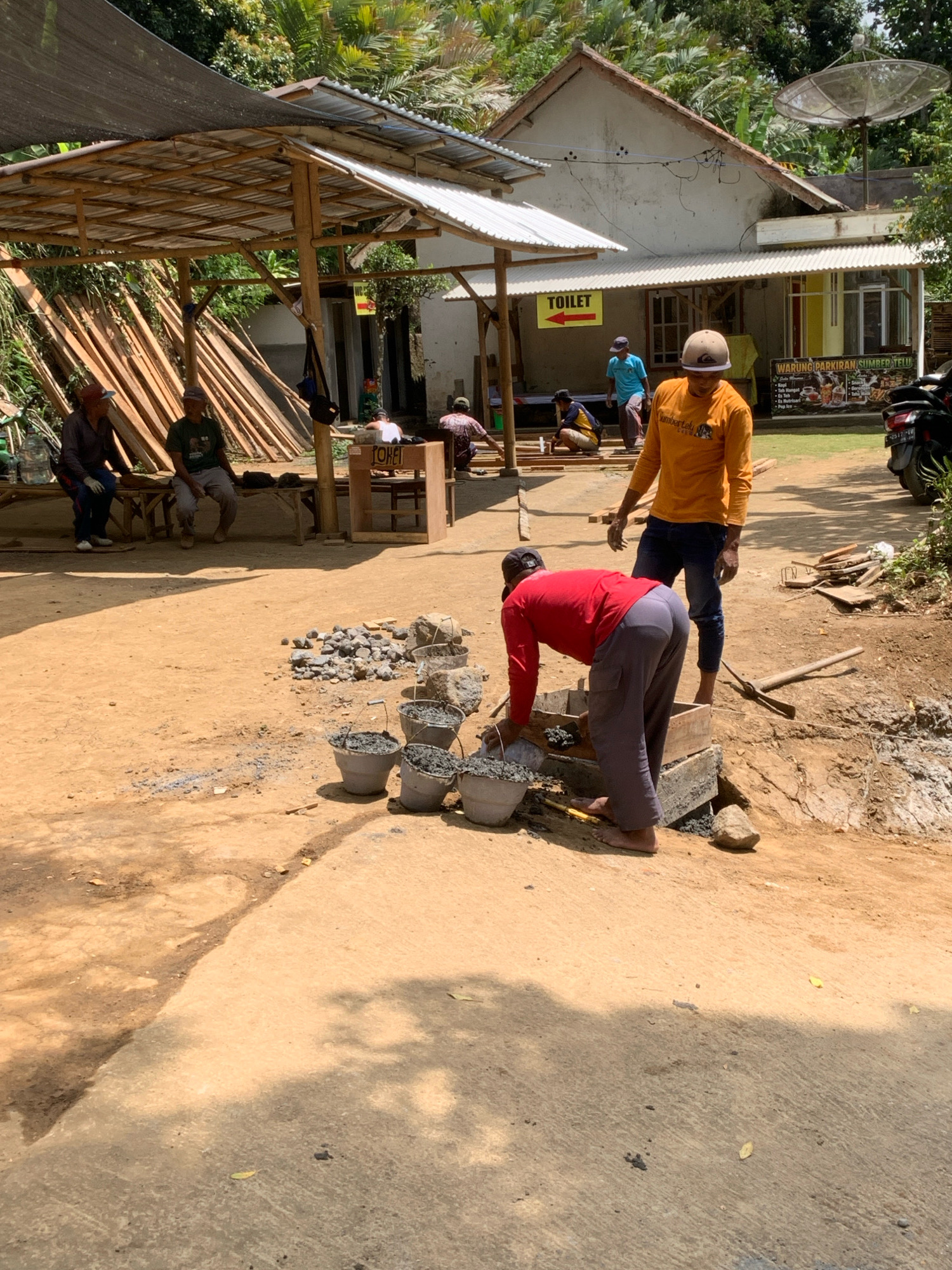 The image depicts an outdoor construction site in Pronojiwo, Indonesia. In the foreground, two men are engaged in construction work. One man, wearing a red long-sleeved shirt and grey pants, is bending over a wooden structure, appearing to scoop material from a bucket. The other man, wearing an orange long-sleeved shirt and blue jeans, stands nearby, looking towards the first man. Several buckets, some filled with grey material, are arranged on the ground around them, along with piles of rocks and a shovel.
In the background, a building with a corrugated metal roof is visible, with a sign that reads "TOILET" with an arrow pointing left. A satellite dish is mounted on the roof of the building. To the left of the building, a shaded structure made of bamboo and corrugated metal provides cover, with stacks of lumber leaning against it. Several people are sitting on a bench under this structure, observing the work or resting. A motorbike is parked to the right of the building. The ground is a mixture of dirt and concrete, with scattered debris and shadows cast by the surrounding trees. The environment suggests a rural or semi-rural setting with lush vegetation in the background.