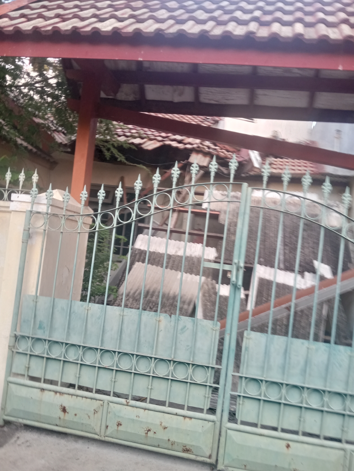 The image captures a scene in Surabaya, Indonesia, depicting a pale green metal gate with intricate circular designs and vertical bars, showing signs of rust and weathering. The gate is partially open, revealing a damaged structure behind it, possibly a house or building, with a tiled roof and visible debris. A concrete wall with decorative finials stands to the left of the gate, and overgrown vegetation peeks through the gaps. The overall impression is one of disrepair and neglect, suggesting a location that has experienced some form of damage or abandonment.