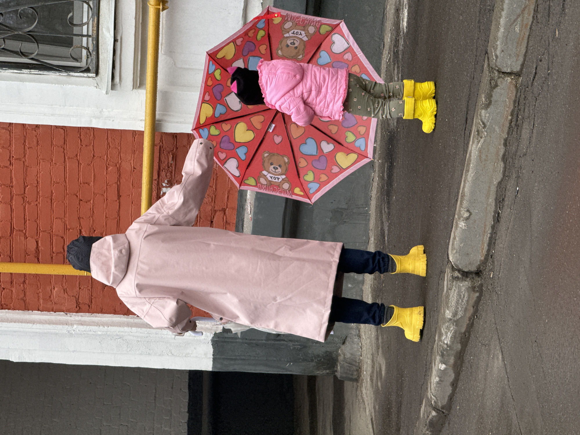 A person in a pink raincoat and yellow rubber boots holds a red umbrella with cartoon bears and hearts. On top of the umbrella, there's a pink jacket and some clothing. The person stands on an asphalt surface near a curb. The scene is near a building with red brick and a window. The photograph was taken in Moscow, Russia.