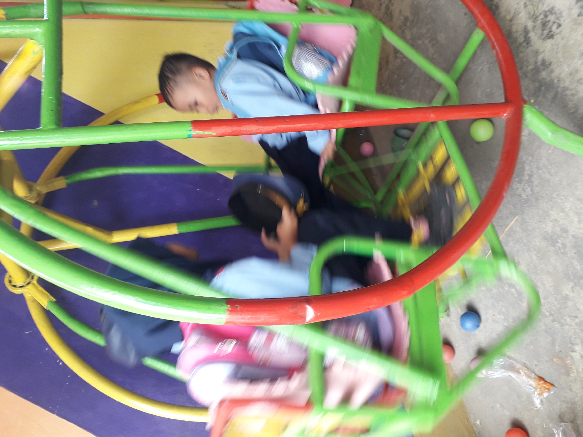 In Cankuang, Indonesia, two children are on a playground swing. One child, with dark hair, is wearing a blue shirt and black pants and is seated on the swing. Another child, blurred by motion, is visible behind them, wearing a dark head covering and a blue top. The swing is green and red with yellow accents and is set against a purple and yellow wall. Several colorful balls are scattered on the ground below.