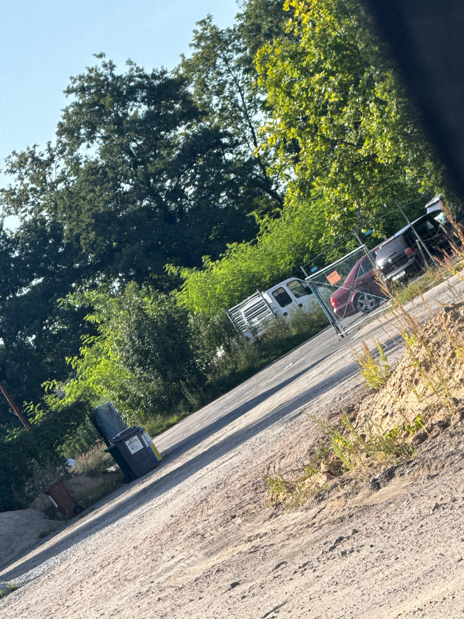The image depicts an outdoor scene, possibly a street or parking area, with a noticeable slant. In the foreground, a dusty, gravelly surface dominates, with tire tracks visible. To the left, two trash bins are present: a dark green one with yellow accents and a smaller, brown bin further back.
In the middle ground, a road or paved area curves away from the viewer. Along this road, a chain-link fence encloses a section where two vehicles are parked. One is a white pickup truck with a metal rack on the back, positioned behind the fence. Inside the fenced area, a red car is visible, and behind it, the rear of a dark-colored SUV or car. The fence has a sign with orange lettering.
The background is filled with lush green foliage, with large trees on the left and denser shrubs and trees on the right. The sky is clear and blue. The overall impression is of an area on the outskirts or in a less developed part of a town, with a mix of vehicles, infrastructure like bins and fencing, and abundant vegetation. The image's composition suggests it was taken from a vehicle or from a slightly elevated position looking down at the scene.
Based on the provided context, this scene is located in Nowa Sól, Poland.