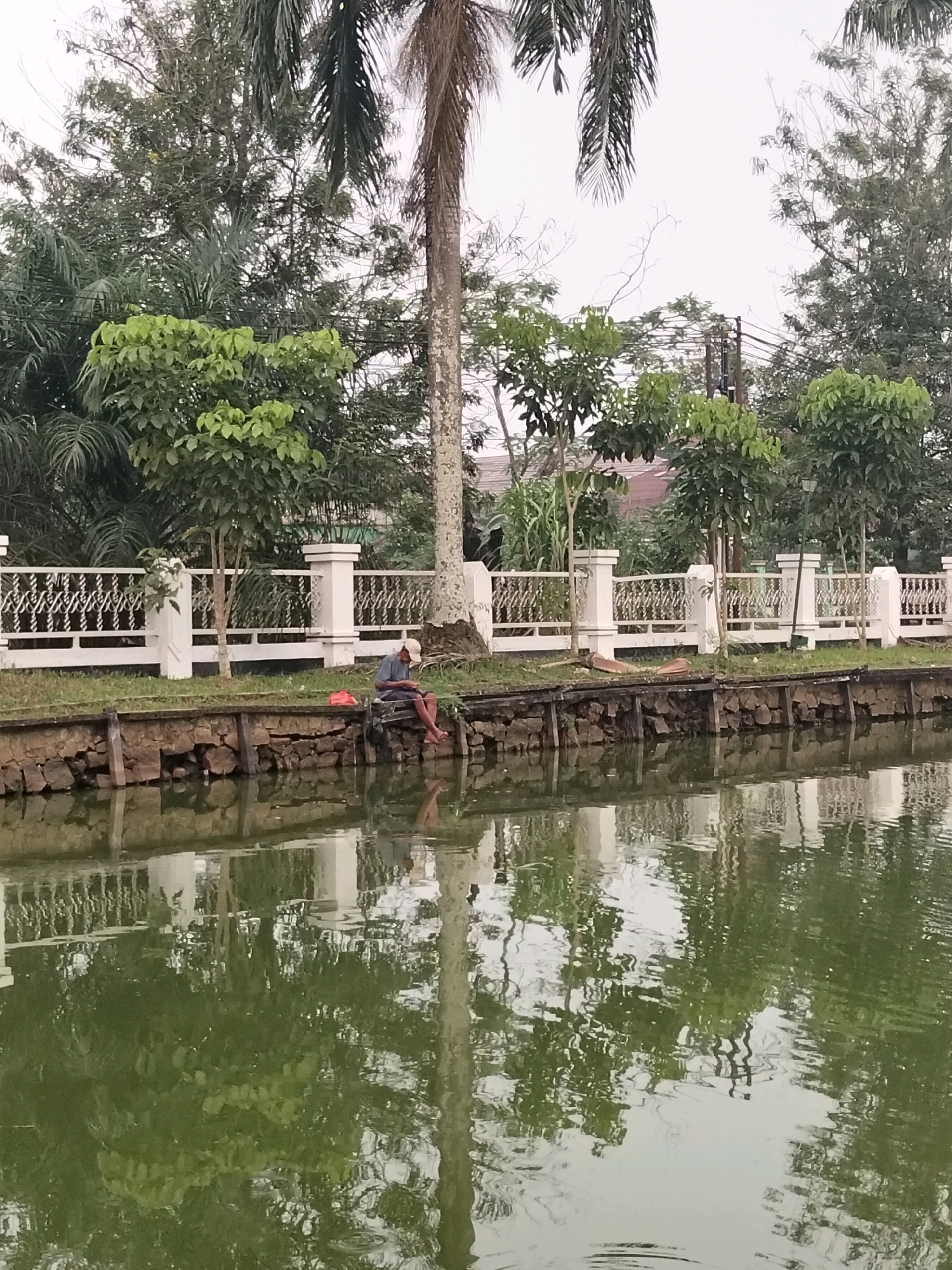 The image depicts a man fishing from the edge of a body of water in Sungai Raya, Indonesia. The man, wearing a blue shirt and a light-colored cap, is seated on a wooden platform by the water, with his legs dangling. He is holding what appears to be a fishing rod and is looking down towards the water. Beside him is a red bag. The water is calm and reflects the surrounding greenery and a white decorative fence. The fence is supported by square pillars and features an intricate metalwork design. Behind the fence, there are various trees, including palm trees, and some buildings are visible in the distance. The ground leading up to the fence is covered in grass. The scene appears to be set in a park or a residential area. The sky is overcast.