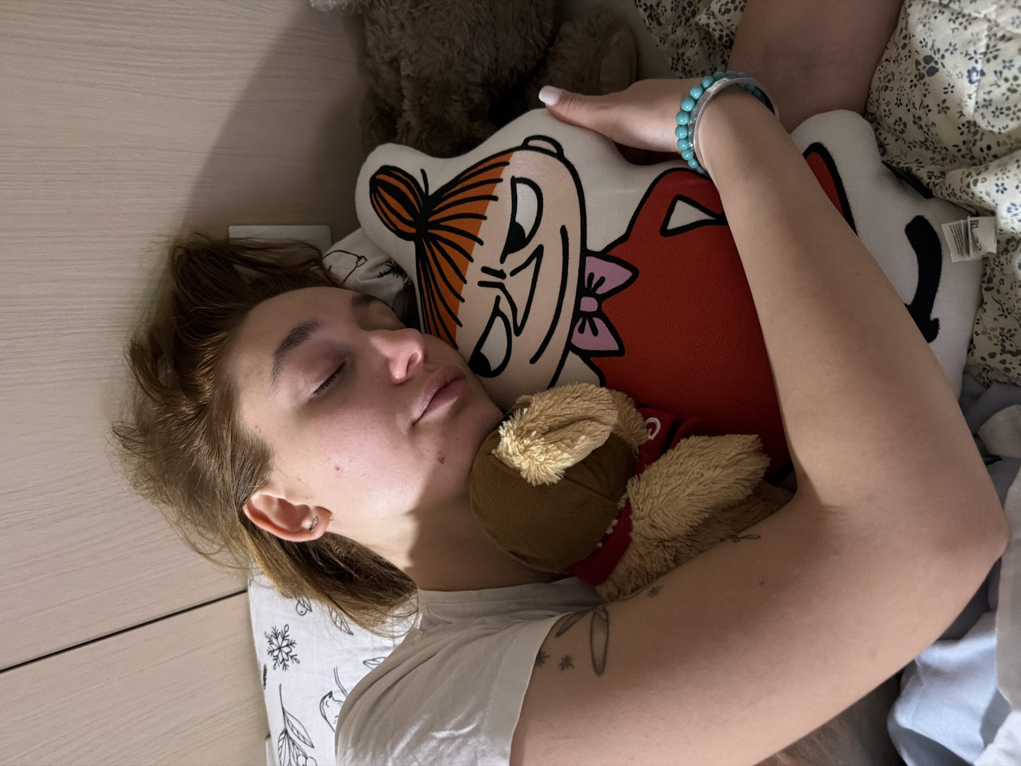 The image captures a top-down view of a young woman resting or asleep in a bed, oriented diagonally from the top left to the bottom right. She is lying on her left side with her eyes closed, suggesting a state of relaxation or sleep. Her hair is light brown or reddish-brown, and she wears a small earring in her visible right ear.
She is dressed in a plain white t-shirt. Her right arm is bent and rests on a white decorative pillow shaped like Little My, a cartoon character from the Moomin series, depicted with orange hair in a bun, a red dress, and a pink bow. Around her right wrist, she wears a light blue or turquoise beaded bracelet. A small, light brown plush teddy bear with a red collar is nestled against her neck and shoulder area. A faint tattoo with what appears to be a plant or abstract design is visible on her left upper arm.
The bed is adorned with a white pillowcase featuring a subtle pattern of snowflakes and other motifs. A floral-patterned duvet or blanket, predominantly blue and white, is partially visible on the right side of the frame. In the background, behind the cartoon pillow, a larger, dark brown plush toy is partially visible. The left side of the image shows a wooden-patterned headboard or wall panel, indicating an indoor setting.
The lighting in the room is dim, suggesting it might be evening, night, or early morning. There is no visible text that is clearly legible, though a small tag with faint markings is present on the back of the cartoon pillow. The overall scene conveys a sense of peaceful rest in a bedroom environment.