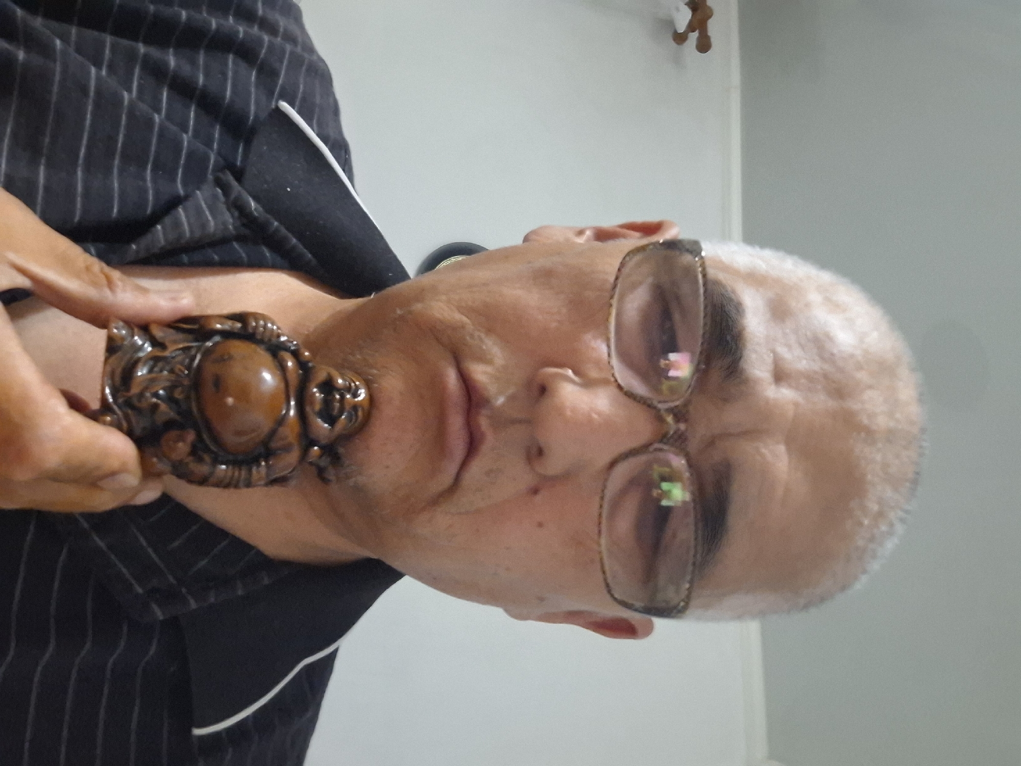 The image shows a close-up, chest-up, selfie-style photo of a middle-aged man with short, grey hair and glasses. He is wearing a black collared shirt with thin white stripes. The man is holding a small, carved wooden figurine of a laughing Buddha in front of his chin with his right hand. The background is a plain, light-colored wall. There is no indication of the location being Almaty, Kazakhstan.