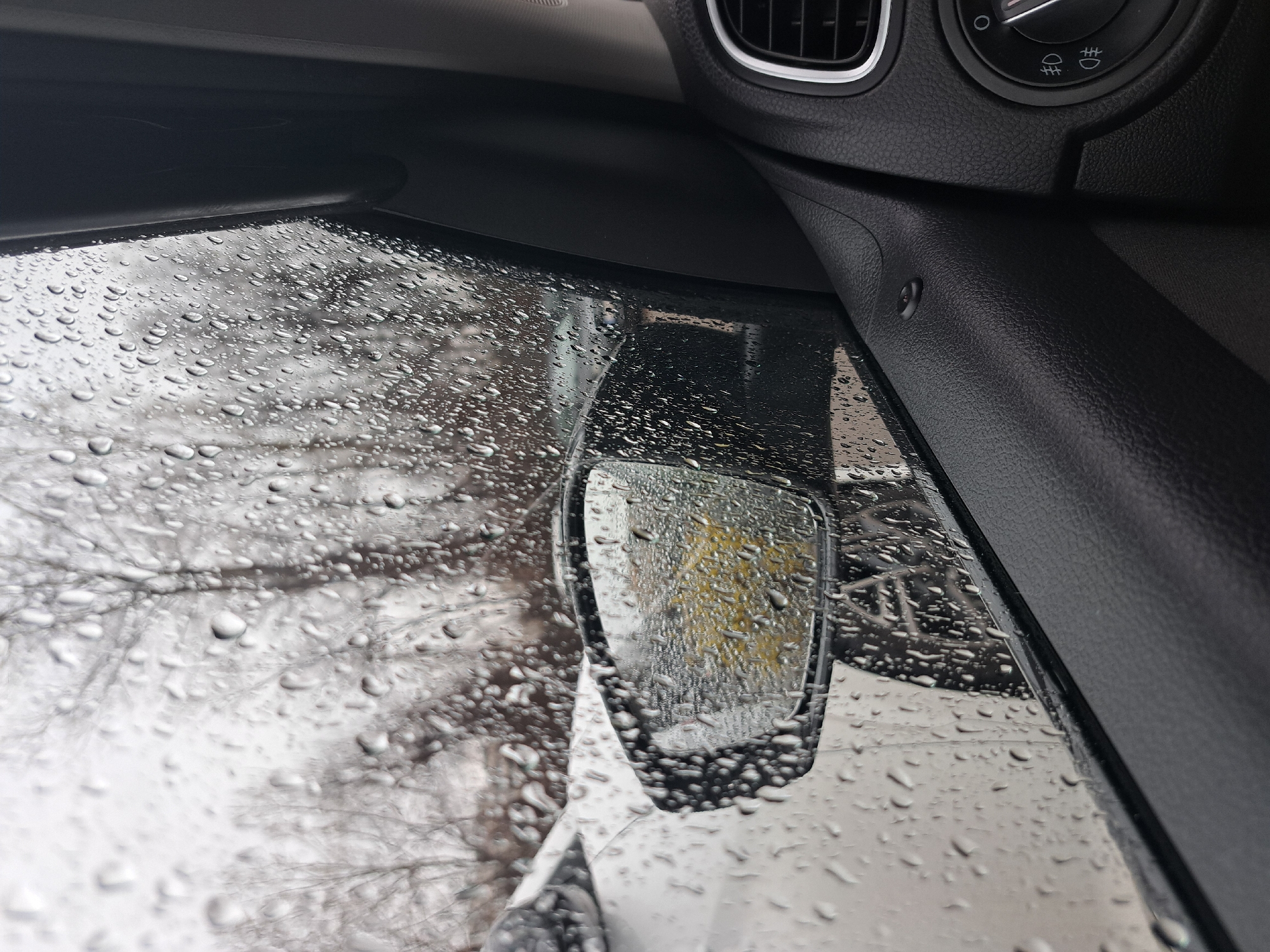 The image is taken from inside a vehicle, likely a car, looking out a rain-splattered side window and side mirror. The window is heavily covered in raindrops obscuring the view, but appears to be a street scene with bare trees. The side mirror provides a distorted reflection of a yellow building or object. The interior of the vehicle is visible, showing a black dashboard, air vent, and light control panel.
Without additional visual context, it is not possible to determine the specific location in Sofia, Bulgaria.