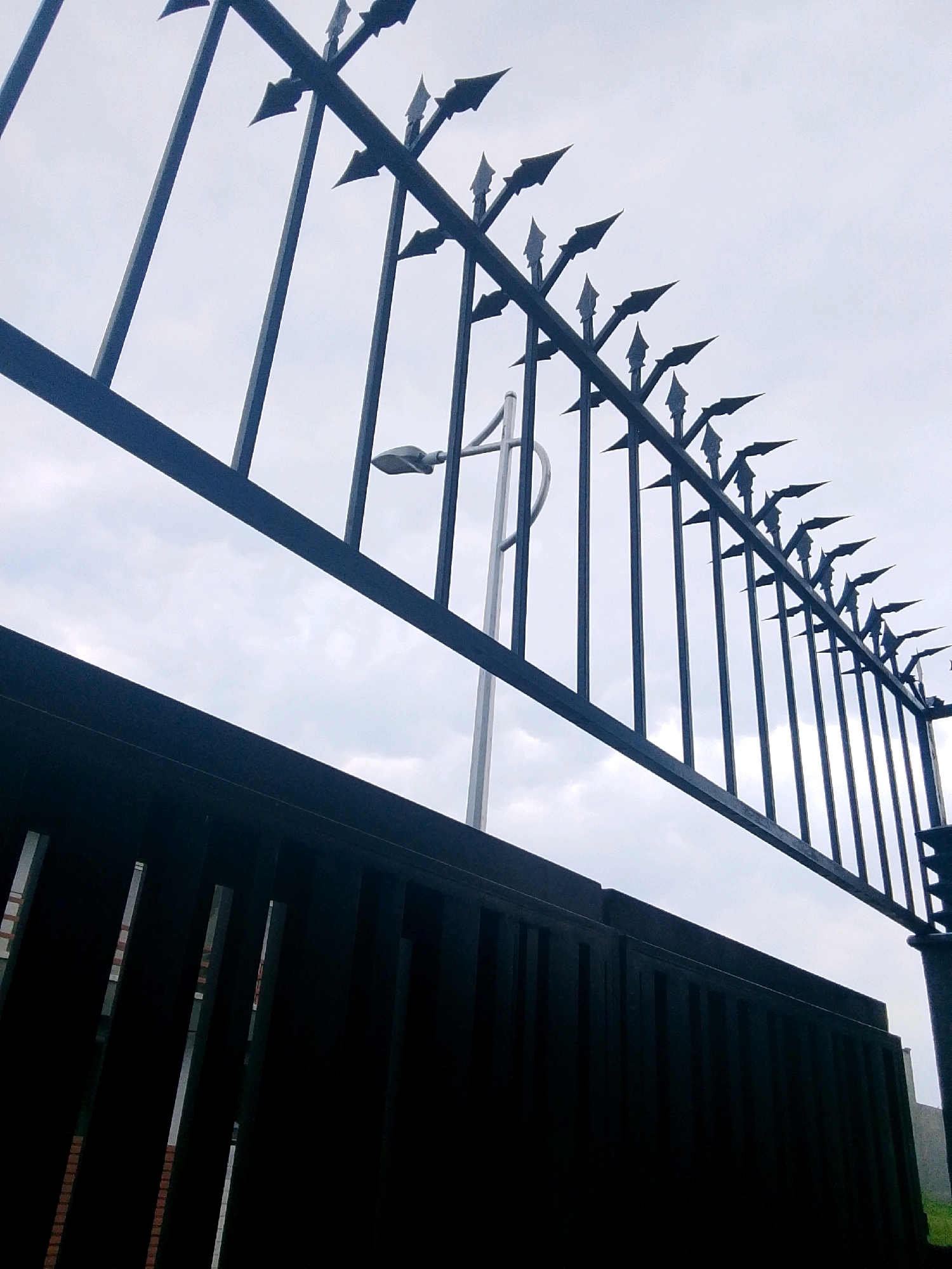 The image depicts a close-up, low-angle view of a dark metal fence with pointed spikes at the top, set against a cloudy sky. In the foreground, a solid dark fence is visible, obscuring the lower portion of the scene. Behind the spiked fence, a modern street lamp with a distinctive shape is mounted on a pole. The overall impression is one of a secure or possibly imposing perimeter. The image appears to have been taken in the city of Taxila, Pakistan, though no specific landmarks are identifiable within the frame to confirm a precise location.