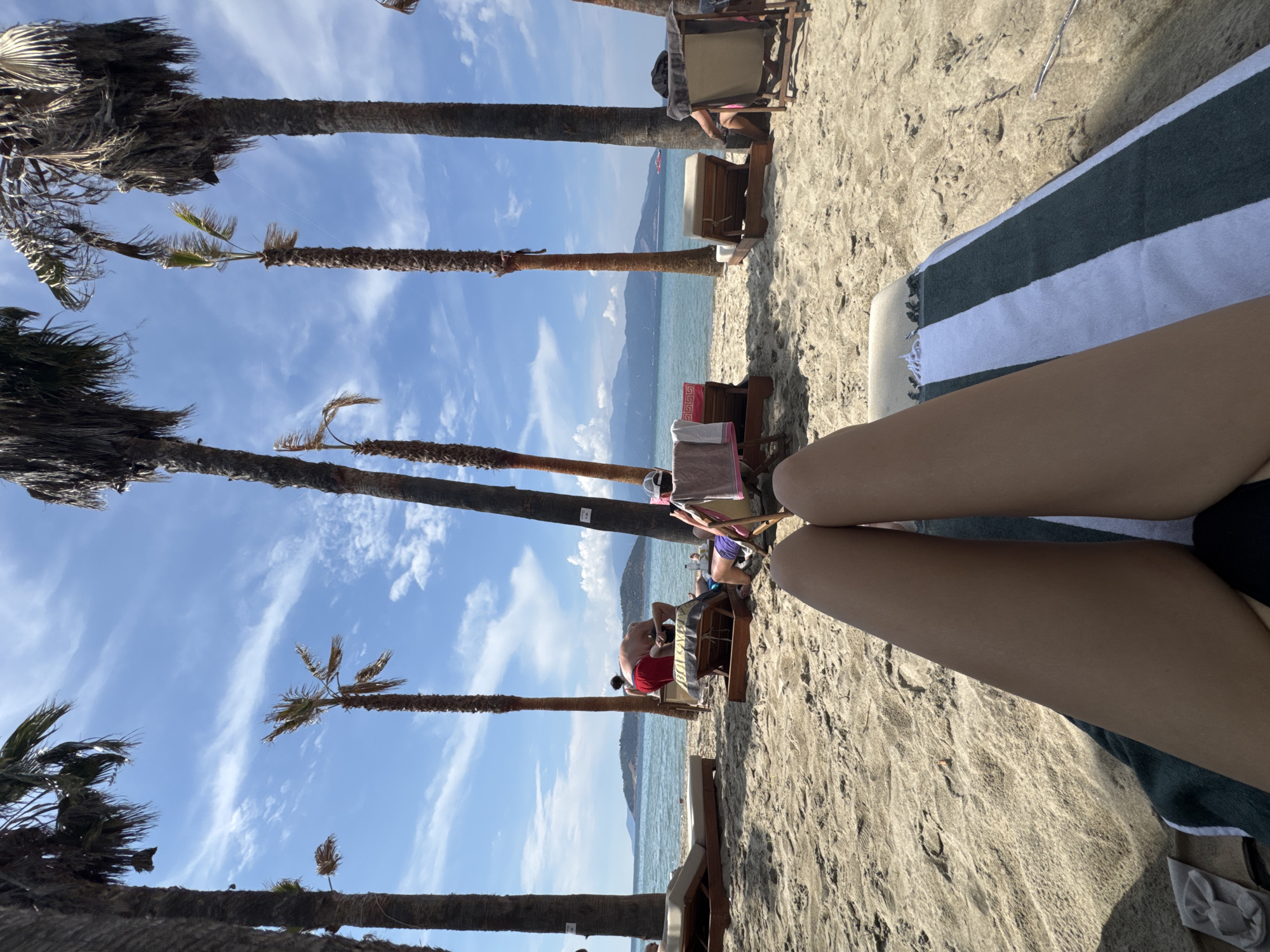 The image depicts a beach scene in Keramoti, Greece, on a sunny day with a bright blue sky and scattered clouds. The foreground features the legs of a person lying on a beach chair, covered by a striped towel. In the mid-ground, several palm trees line the sandy beach, casting shadows. Beach chairs and loungers are visible, some occupied by people relaxing or getting up. One person in red shorts is bending over a chair, while another person in purple shorts sits in a chair. The background shows the tranquil blue sea, with a distant island or peninsula covered in vegetation and mountains. A small red boat is visible on the water in the far distance. The overall atmosphere is one of leisure and relaxation on a tropical-style beach.