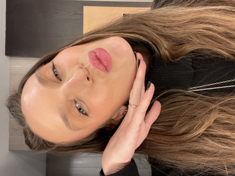 A young woman with light skin and pink lips rests her hand on her cheek, showcasing a gold ring. She has long, light brown hair. Her eyes are open and light-colored, enhanced by dark eyeliner. She's wearing a black turtleneck sweater, and a silver chain is visible around her neck. The background features a neutral-toned surface and a dark, rectangular shape, possibly a piece of furniture or decor. The image is presented at an angle.