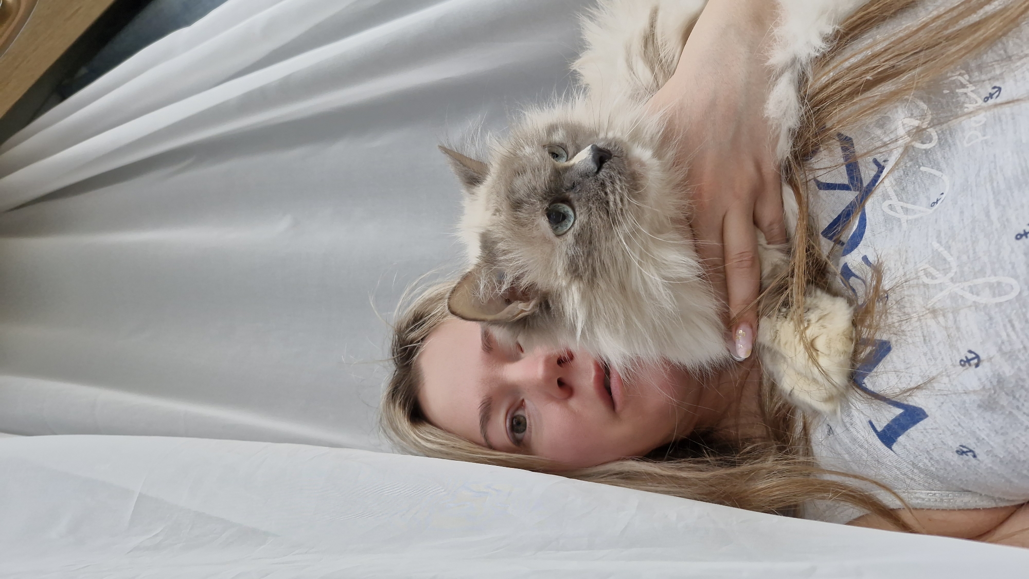 A woman is lying down with a cat on top of her. The woman has blonde hair and is wearing a light gray shirt with blue anchor designs. The cat is a light-colored breed with blue eyes. They are lying on a white surface. There is a wooden bed frame partially visible in the upper left corner. There is no information available that indicates the location is Otchor, Russia.