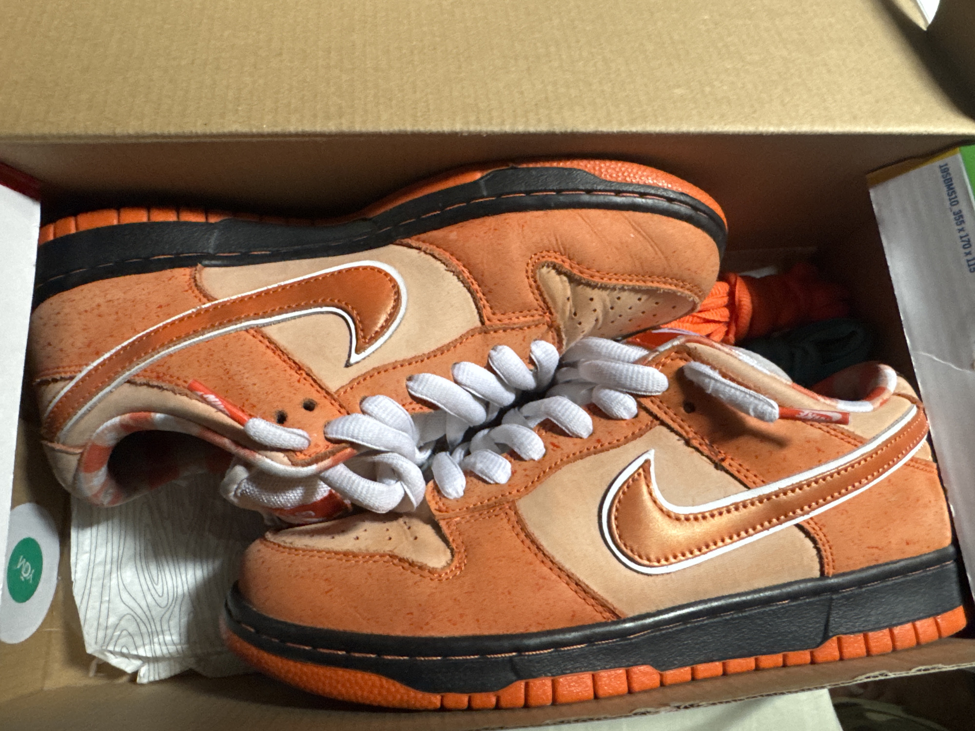 Inside a cardboard box, two orange and tan Nike Dunk Low sneakers with white laces are placed on top of tissue paper. The shoes have orange soles, black midsoles, and orange detailing around the Nike swoosh. Additional orange and green shoelaces are tucked behind the shoes. A green and white circular sticker is visible on the left side of the box, and a green and white informational label is present on the right side of the box. The scene is indoors and well-lit, but the location in Barcelona, Spain is not determinable from the provided visual information.