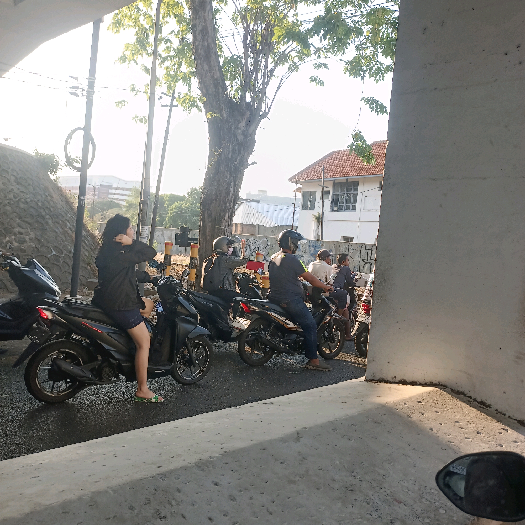 In Surabaya, Indonesia, at what appears to be an intersection on a sunny day, a group of people are on motorcycles, possibly waiting for a traffic signal. A woman in a black jacket and blue shorts sits on a black motorcycle, adjusting her hair. Behind her, several other motorcycles are lined up. One person is wearing a helmet and holding another helmet. Further back, other individuals are seated on motorcycles, some with passengers. The environment includes a concrete structure, a large tree with green leaves, utility poles with wires, and a building with a red tile roof in the background. The road surface is asphalt, and sunlight casts shadows.