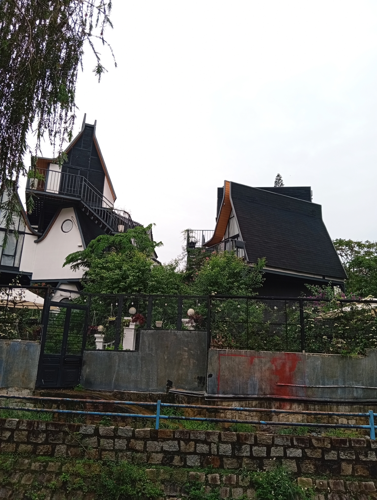 Two distinctive houses with unique architectural designs are situated on a slope, separated from the street by a wall and fence. The house on the left is white with black framing and a prominent staircase leading to an upper level with a balcony. The right house features a dark roof, possibly black shingles, and a contrasting wooden frame. Lush greenery and flower bushes surround the houses and grow along the fence. Below the fence, a stone wall is visible, and a blue metal railing runs along its top. The scene is set in Da Lat, Vietnam, known for its unusual architecture. The houses are of unusual design, with sharp lines and angles. Overcast lighting permeates the scene.