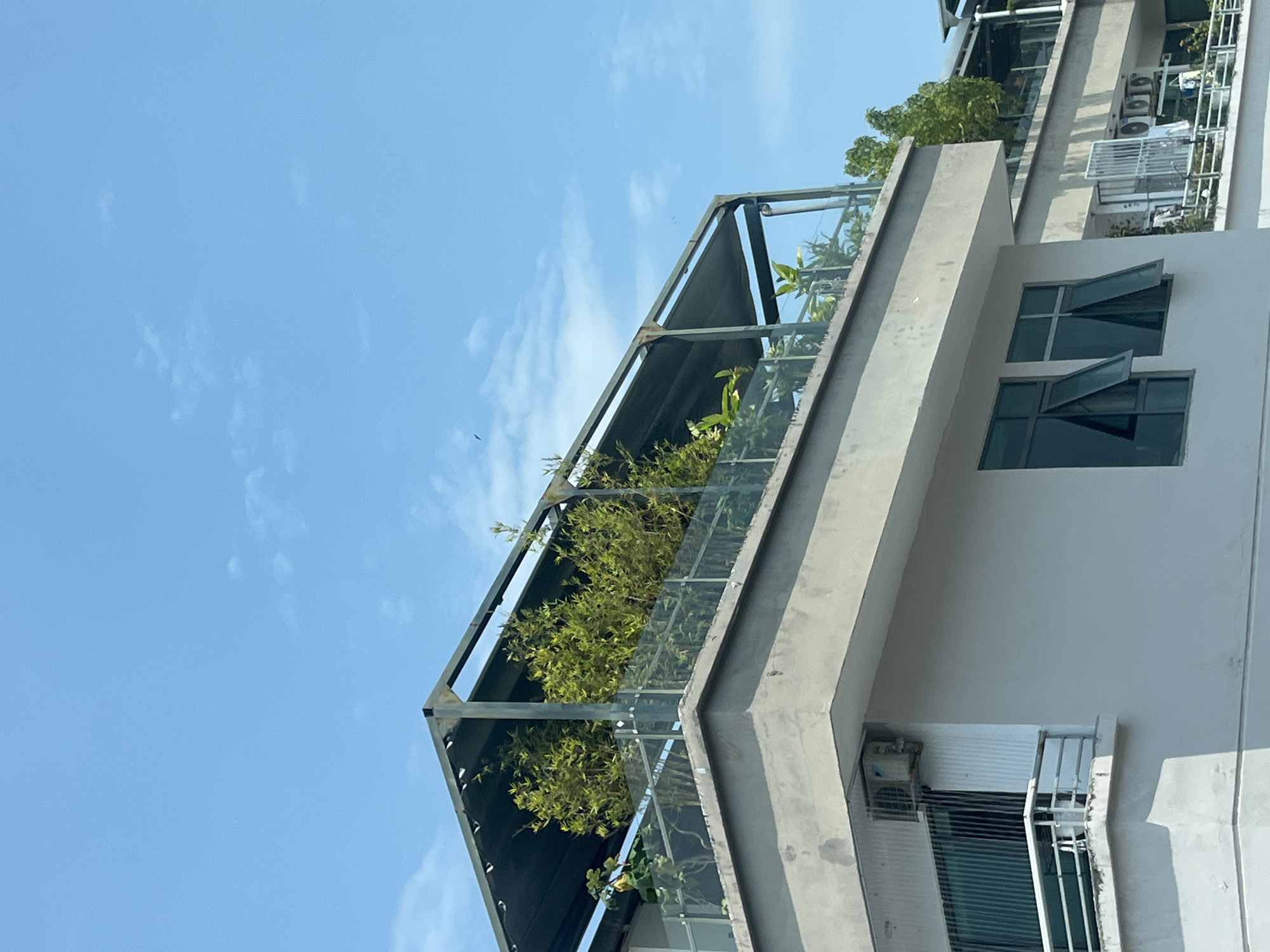 The image shows the exterior of a modern building, likely a residential apartment, in Bac Nha Trang Ward, Vietnam. The building is painted in a light color. Two windows are partially opened on the building's facade. An air conditioning unit is visible on the wall. A balcony on the upper floor is enclosed with a metal frame and glass panels and contains greenery, including plants and small trees. The sky is clear and blue, with a few clouds.