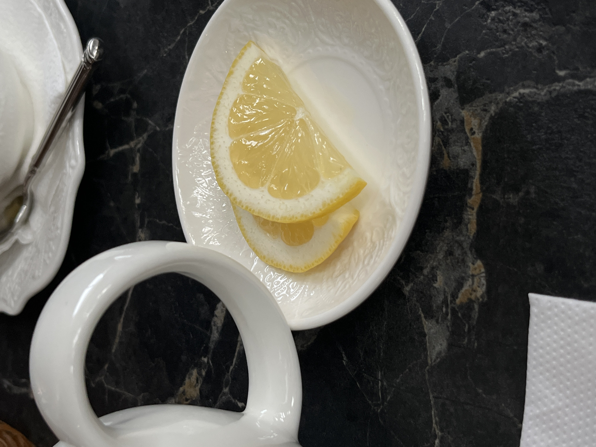 The image shows a close-up of a table setting in what may be a restaurant or cafe. The table has a dark, possibly marble-like, surface. Two lemon slices are placed in a small white saucer with a decorative, embossed pattern. To the left is part of a white teacup and a small silver spoon on a separate, smaller saucer. In the bottom left corner, a portion of a white teapot handle is visible. In the lower right corner, there's a white paper napkin. There are no people visible. The location is not discernible from the image, though it was specified to be in Sochi, Russia.