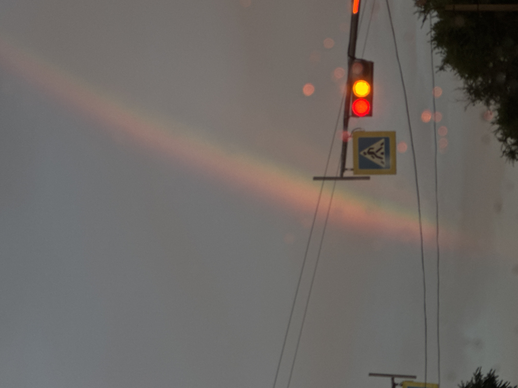 A faint rainbow arcs across a cloudy, gray sky. In the foreground, a traffic signal displays a red and yellow light. Below the traffic signal, a pedestrian crossing sign is visible. Power lines crisscross the scene. In the lower right corner, the tops of dark green trees are partially visible. The overall atmosphere is muted and overcast, suggesting recent or impending rain.