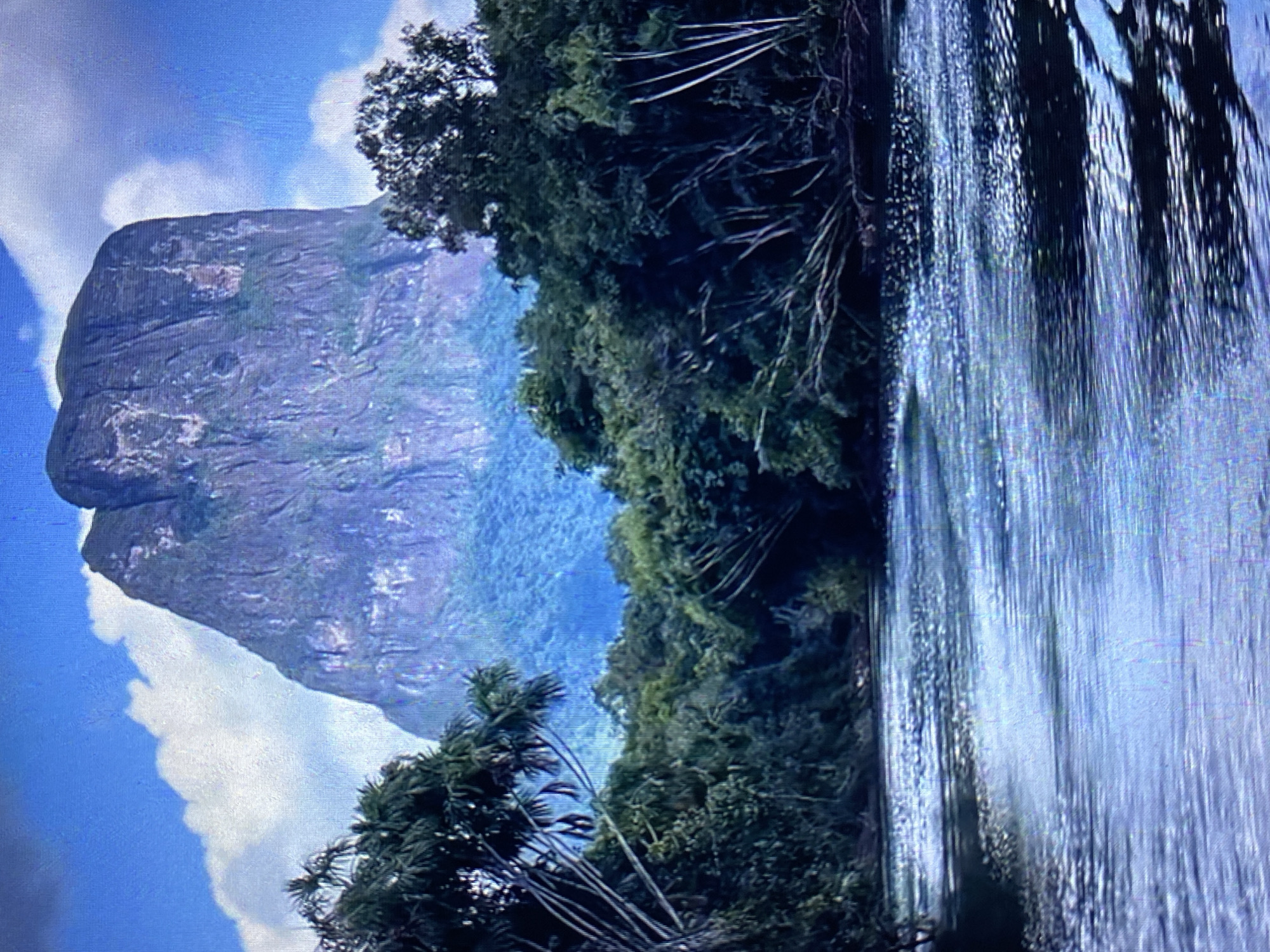 The image shows a natural landscape, likely near Tinaquillo, Venezuela. A large, rocky mountain dominates the left side of the frame, with scattered clouds in the blue sky around it. Dense green vegetation, including trees and possible jungle growth, covers a large portion of the midground and right side of the image. The water body is in the foreground. The image is a landscape shot, potentially depicting a remote or natural area of Tinaquillo.