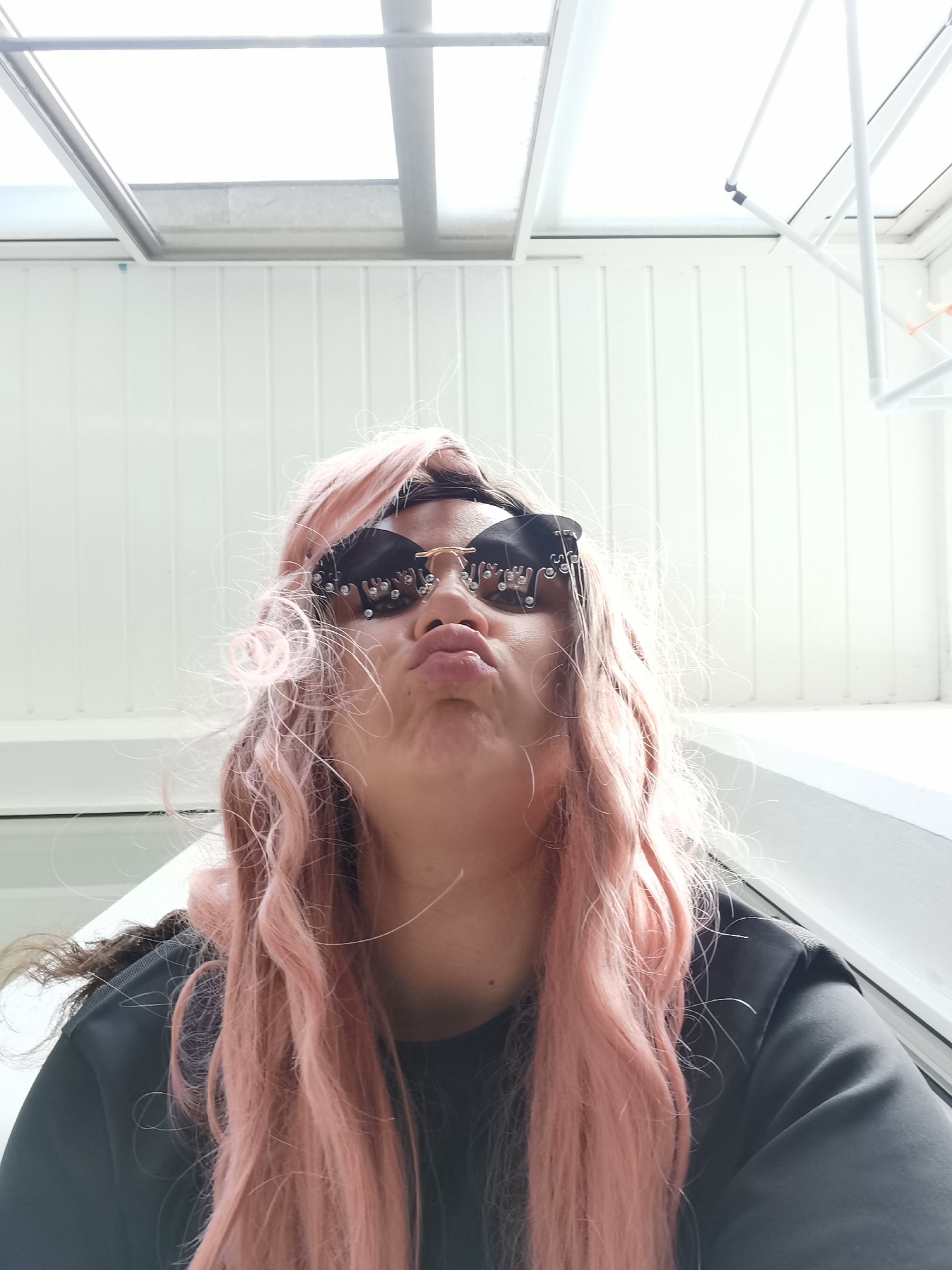 A person with pink, wavy hair and wearing black sunglasses with crystal embellishments is puckering their lips in a selfie taken from a low angle. The person is wearing a black top. Behind the person is a white wall with vertical lines and a window above with a white grid pattern. The location appears to be indoors with bright, natural lighting, possibly in Chaikovsky, Russia.