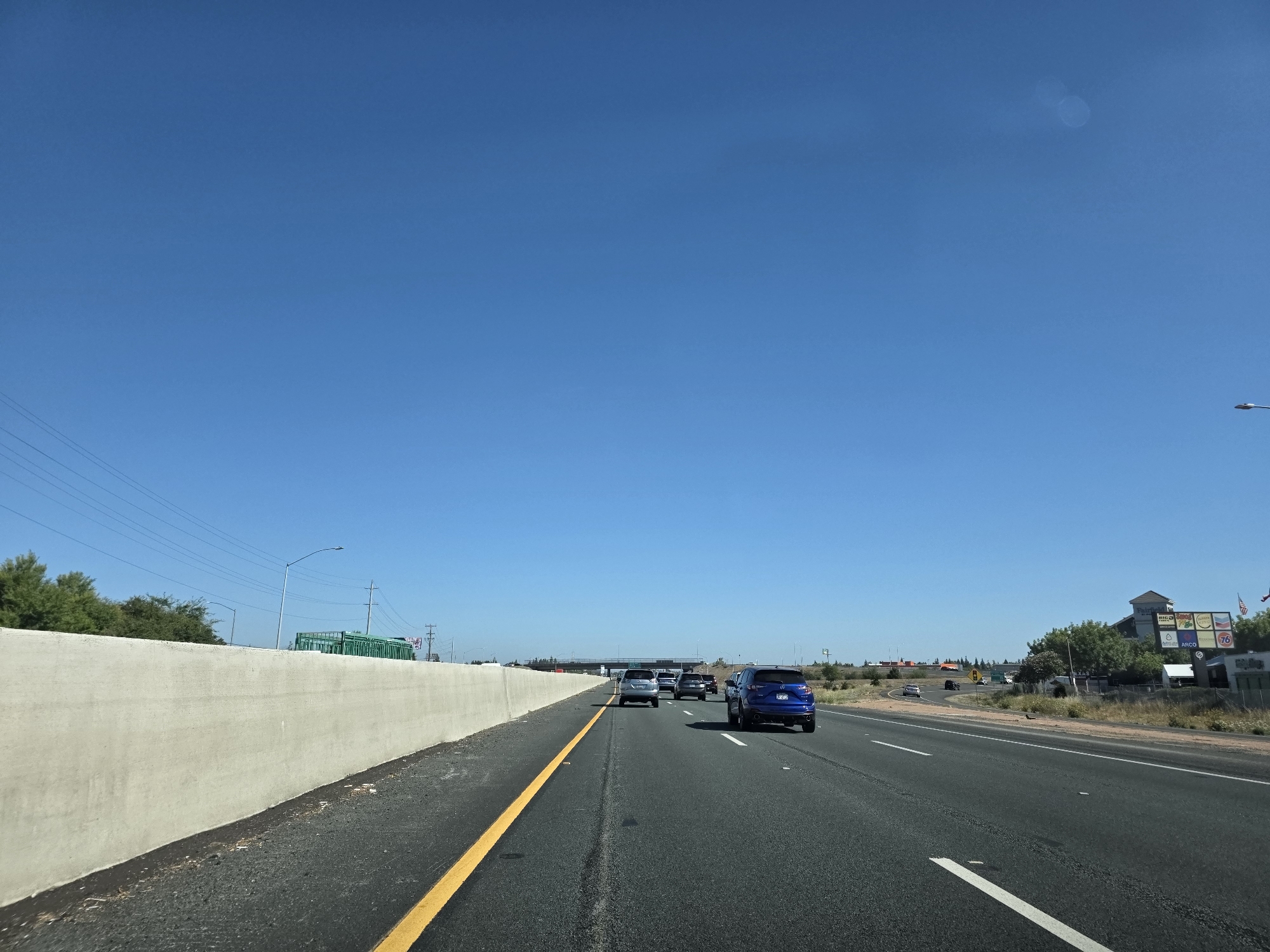 On a clear, sunny day in Elk Grove, California, USA, a blue SUV travels down a multi-lane highway. The vehicle is in the right-hand lane, moving alongside other cars. To the left of the highway is a concrete barrier, and beyond that, trees and power lines. On the right side of the highway, there are businesses and signs, including a 76 gas station. The sky is a vibrant blue with no clouds.