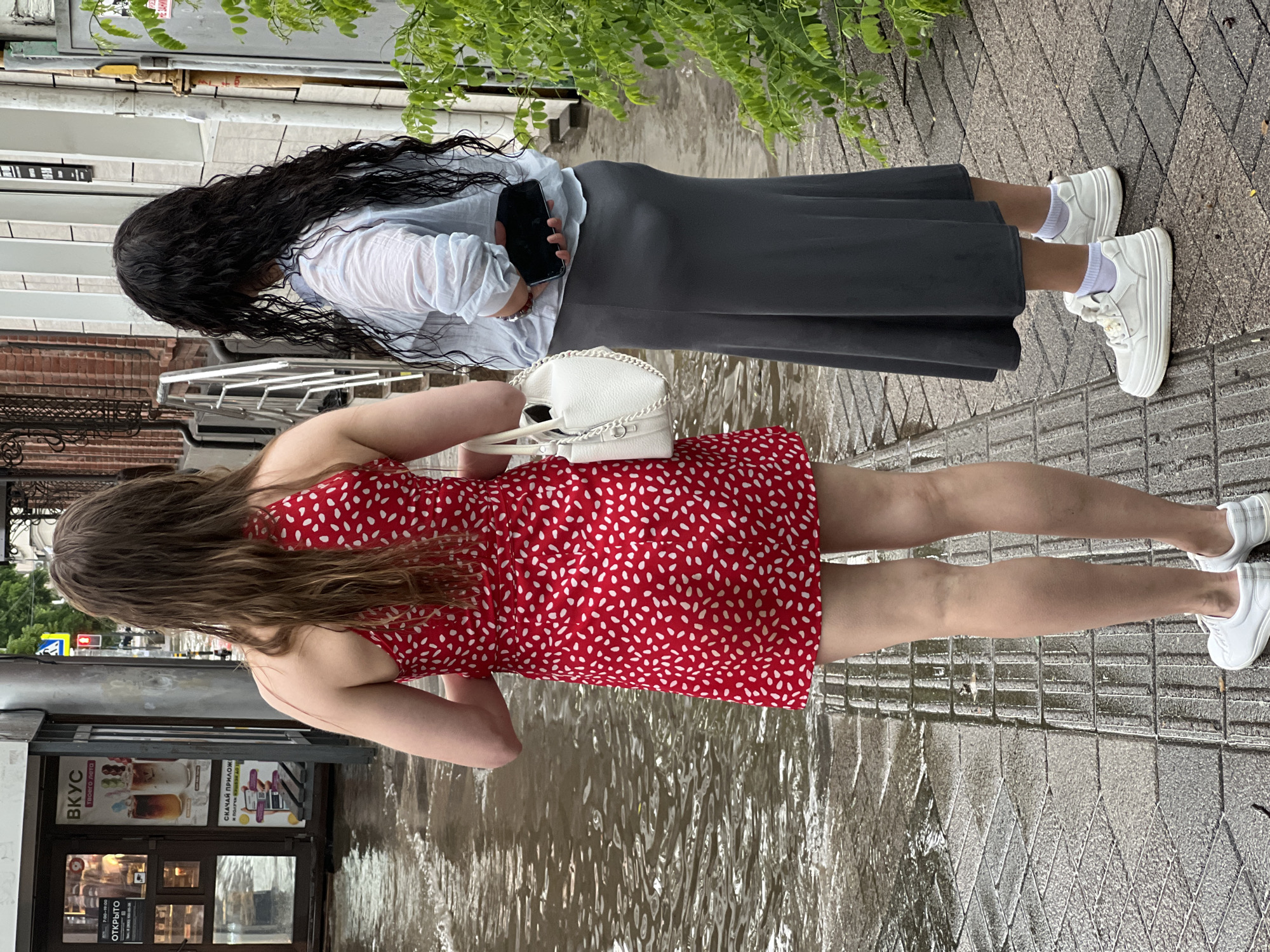 Two women stand on a flooded sidewalk in Krasnodar, Russia. The woman on the left wears a red dress with white polka dots and carries a white handbag. The woman on the right wears a light blue shirt and a long grey skirt and holds a dark smartphone in her hand. The sidewalk is paved with grey tiles and is submerged in shallow, murky water, indicating recent heavy rainfall or poor drainage. Behind the women, a storefront with a glass door and advertisements is visible. The environment suggests an urban setting, possibly a commercial street, experiencing inclement weather.