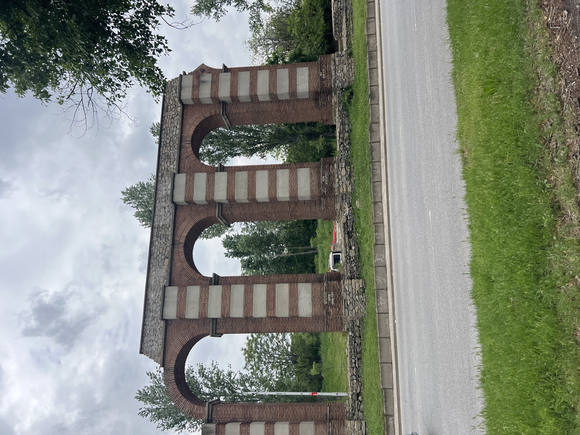 The image shows the Three Arches structure located in Plovdiv, Bulgaria. The structure is constructed of brick and stone, with three prominent arches. It stands adjacent to a road, with greenery and trees visible in the background and foreground. Overcast skies are present above.