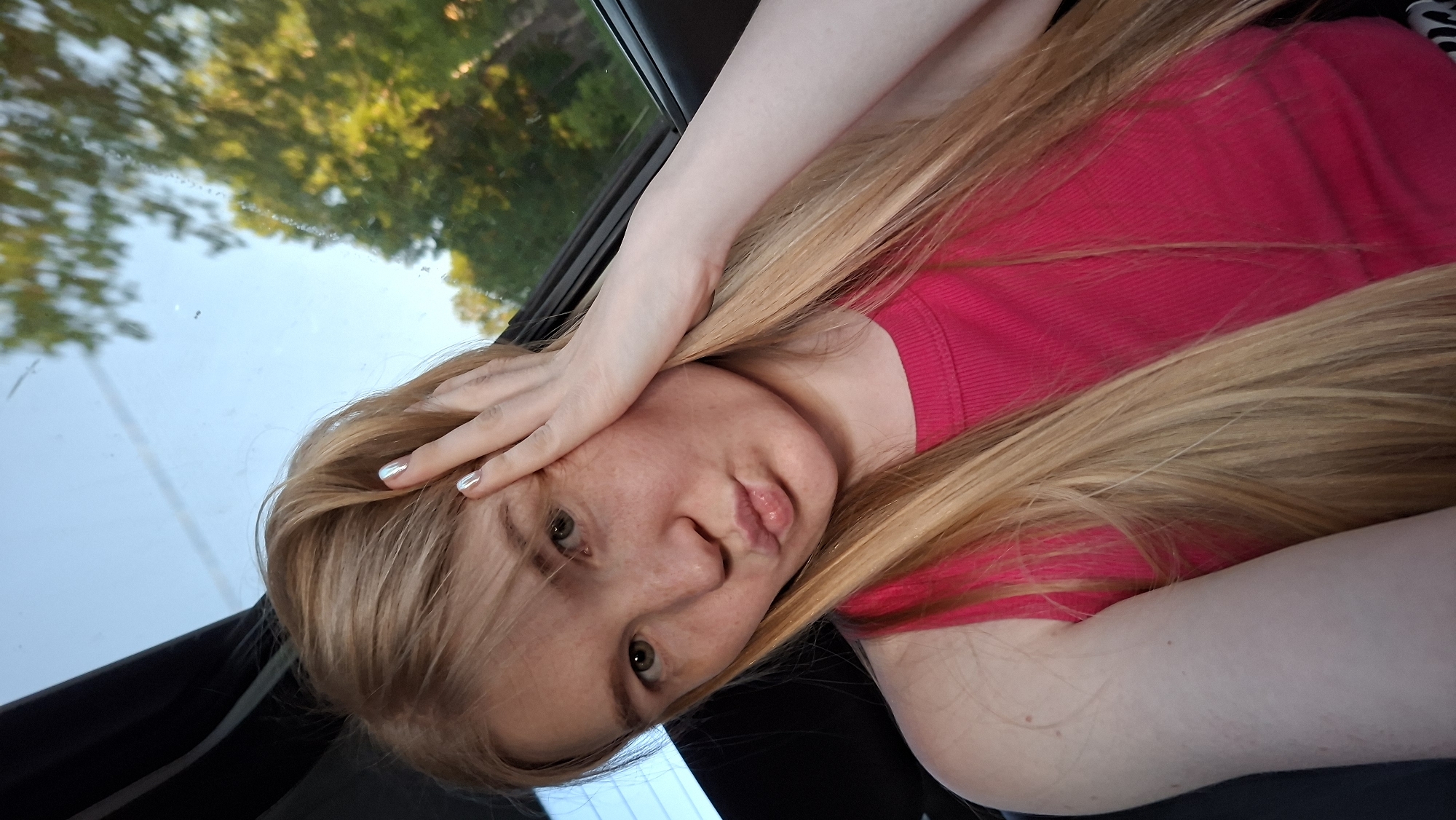 A young, light-skinned woman with strawberry blonde hair and green eyes is looking into the camera. She is wearing a pink shirt and her hair is neatly combed, but there is also some hair covering her forehead. She has one hand on her forehead and the other at her side. She is in a car looking out through the windshield. A light blue sky and the tops of green trees can be seen through the windshield. The location is uncertain.