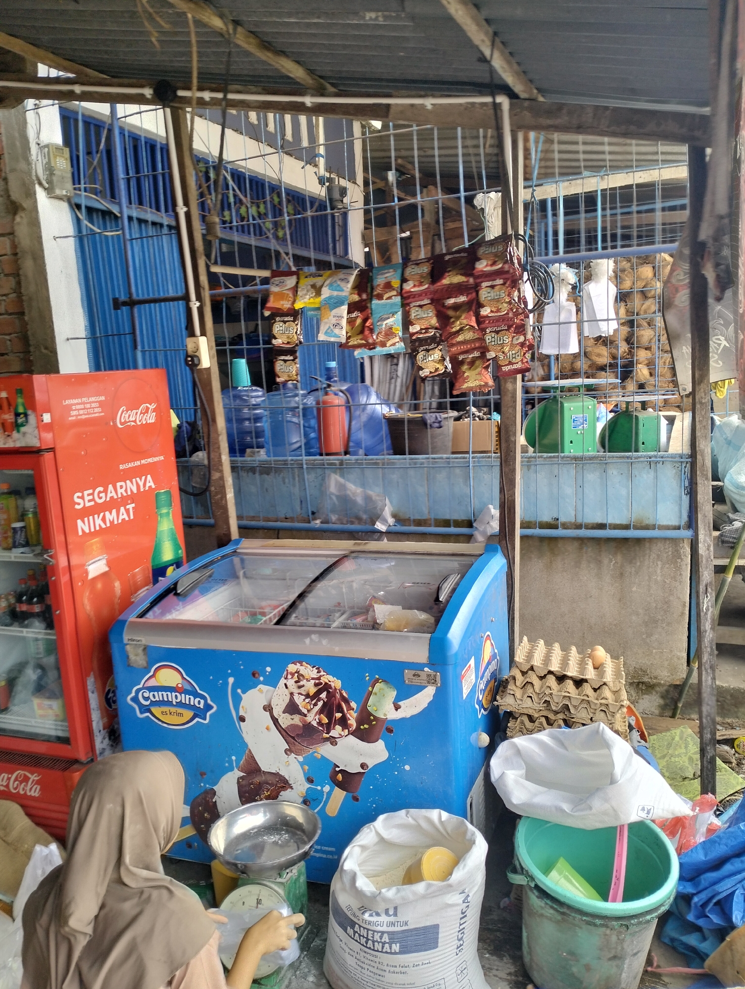 The image depicts an outdoor scene in Serdang Jaya, Indonesia, likely a small shop or market stall. In the foreground, a person wearing a beige hijab is operating a mechanical scale, with a stainless steel bowl placed on it. Beside them are two large white sacks, one partially open and revealing a yellow cylindrical object inside, and the other with visible Indonesian text. To the right, a green plastic bucket contains some items, including a pink rod.

Dominating the center of the image is a blue chest freezer with the "Campina es krim" (ice cream) logo prominently displayed, featuring illustrations of ice cream cones. Behind the freezer, a red Coca-Cola branded refrigerator stands, showing various bottled beverages inside. Above the freezer and refrigerator, a wire mesh fence encloses an area where numerous bags of snacks are hanging, with some brands like "Garuda Plus" visible.

Further back, there are stacks of egg cartons, some containing eggs, and several large blue water jugs. The environment appears to be a slightly cluttered, open-air setup with a corrugated metal roof supported by wooden beams overhead. A brick wall is visible on the left side of the frame. The overall impression is that of a humble, functional retail space catering to local needs.