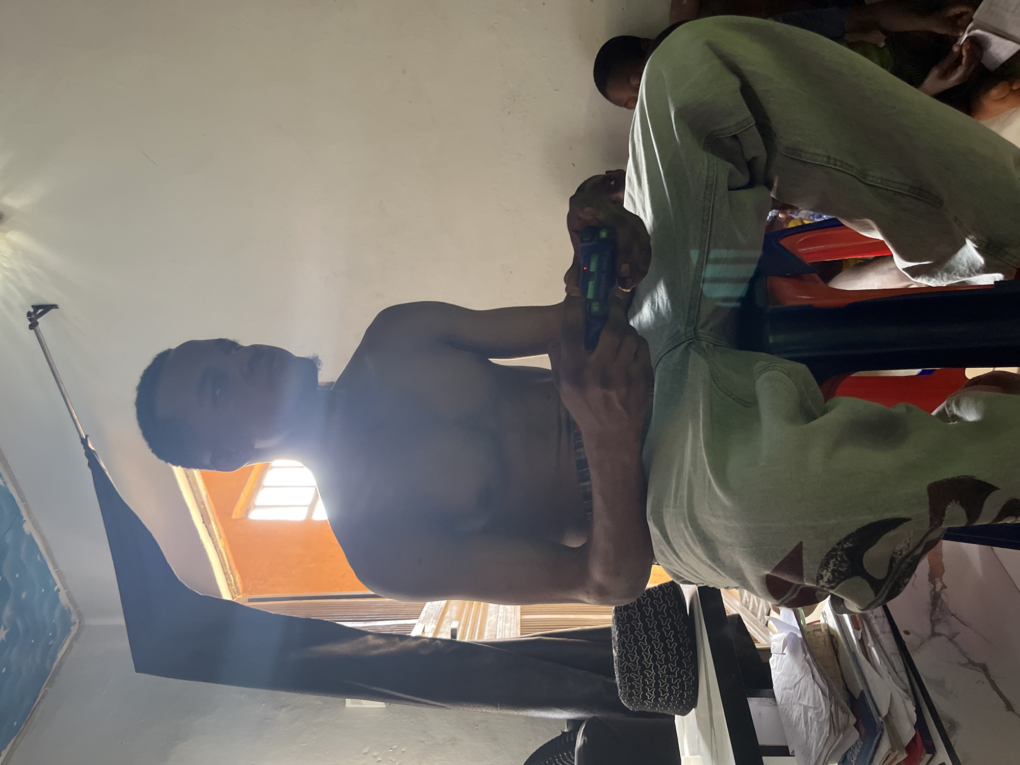 The media shows a group of people indoors, possibly in a room with bright lighting from a window. The perspective is low and tilted, giving an unusual viewpoint. The primary subject is a shirtless individual holding a toy gun, with another person visible in the background. The individual holding the gun is wearing light-colored pants with a patterned design. They appear to be engaged in play or a game. The room's setting is suggested by a window with a view of an orange-colored wall and interior elements like furniture and papers. The overall mood seems casual and playful, despite the potentially provocative object in hand. There is no visible text.
