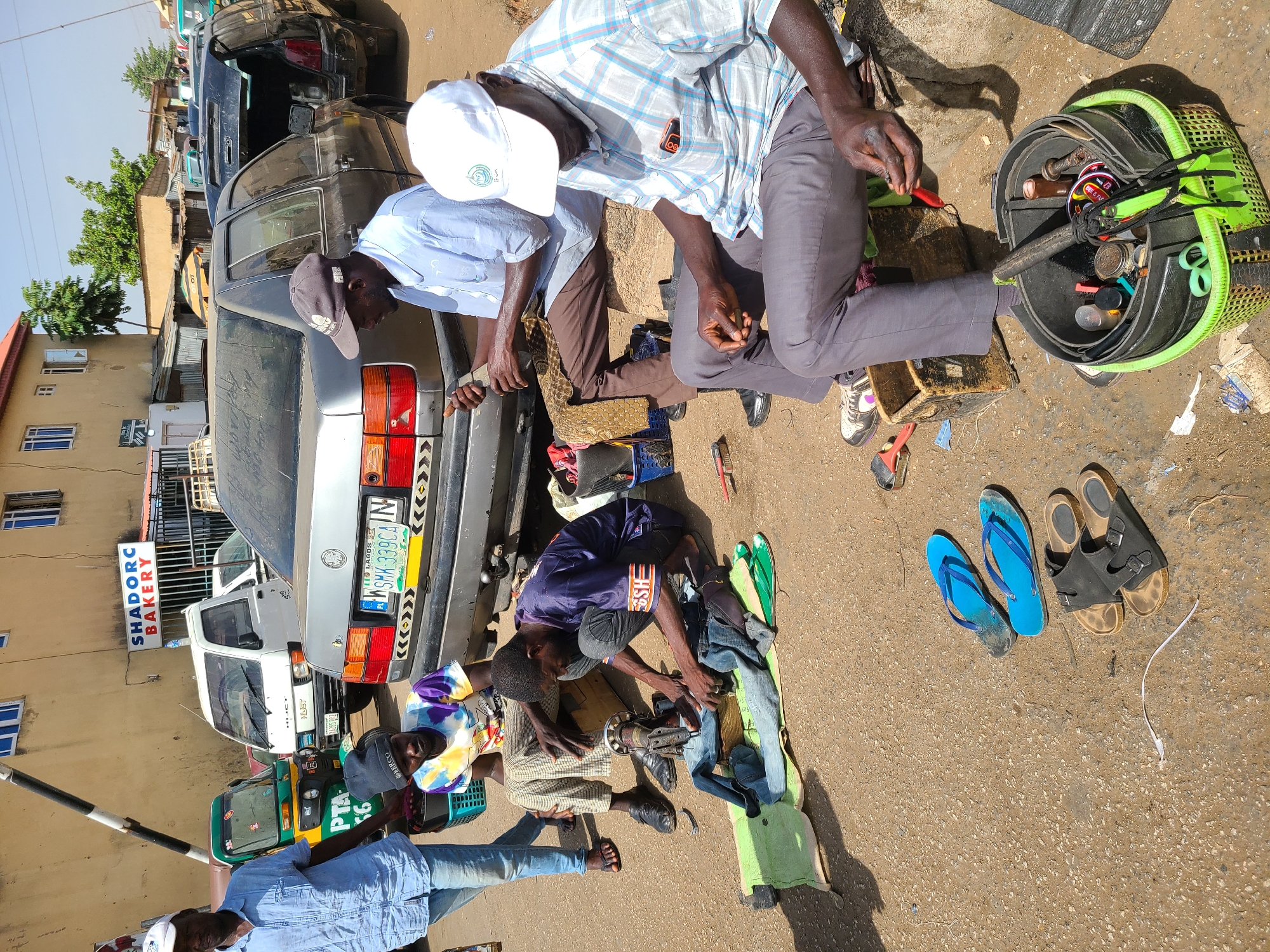 This outdoor image captures a bustling street scene in Jos, Nigeria, under bright daylight conditions, suggesting mid-morning or early afternoon. The primary activity appears to be street-side shoe repair or craftwork, involving several men.
In the foreground, a man in a light blue plaid shirt and dark trousers, wearing a white cap, is squatting and actively working with a red-handled tool. Beside him, a green plastic basket is filled with various tools, such as scissors, pliers, and bottles. Another man, dressed in a light blue long-sleeved shirt and a dark baseball cap, is seated nearby, seemingly observing or assisting. Further into the scene, a man in a blue and purple patterned shirt is seated on the ground, operating a small manual sewing machine on a green mat, with fabric or clothing items arranged next to him. A fourth man, partially visible on the left, wears a light blue denim-like shirt and a white cap.
The ground is unpaved and dusty. Several pairs of sandals and flip-flops, including bright blue and black ones, are scattered around, indicating the nature of the craft.
The background features multiple vehicles. A silver/grey car, displaying a Nigerian license plate from Lagos ("WSMK-339CA"), is parked with its rear visible. Behind it, a white mini-truck is parked, and further left, a green and yellow tricycle, common in Nigeria, is seen with "PTA 56" and "BERCO" written on it. Buildings line the street, including a light brown structure with blue-framed windows and a blue sign reading "SHADORC BAKERY." Sparse trees and vegetation are also present in the background. The men appear focused and diligent in their work, contributing to an industrious street atmosphere. Visible text includes "HUST" partially on one shirt, and "SH" and "2023" on another.